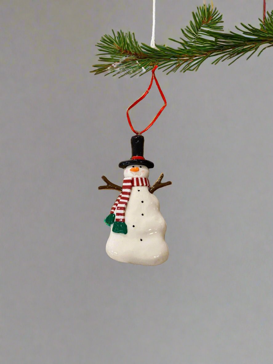 10.8cm small white snowman ornament, wearing a black hat with a red hat band, brown sticks for arms, a red &amp; white striped scarf with a green fringe, and with black dots down his chest and an orange nose, hanging from a tree branch