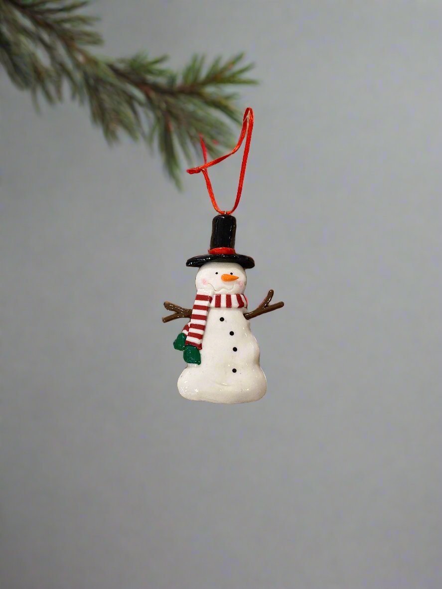 10.2cm white snowman ornament, wearing a black hat with a red hat band, brown sticks for arms, a red &amp; white striped scarf with a green fringe, and with black dots down his chest and an orange nose, hanging from a tree branch.