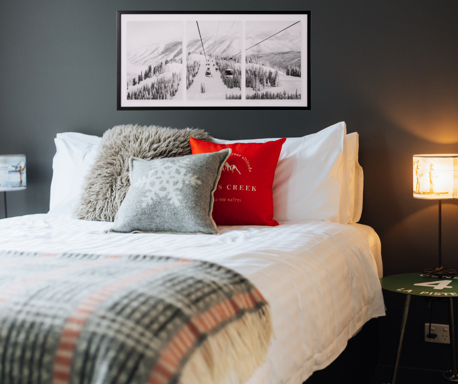 A stunning black and white triptych framed photograph of gondolas ascending a snow covered mountain in a black frame. A triptych is an image in 3 sections. Print show against a dark grey wall, above a large bed with white linen, grey, beige and red cushions and a black, grey, red and white blanket, green ski pole legged side tables and skier lamps with 1 lamp lit.