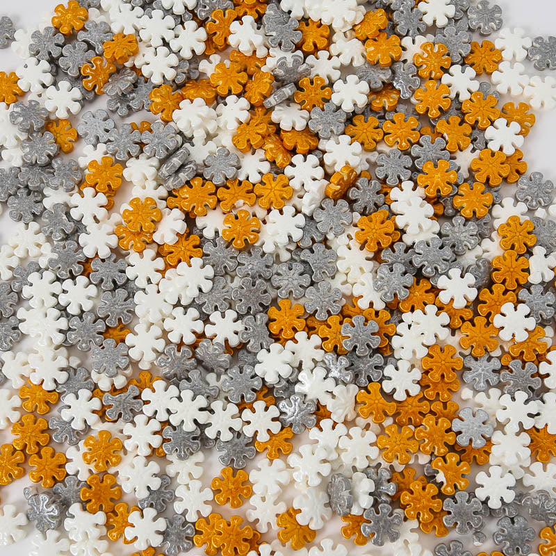 Image of white, silver & gold snowflake shaped sprinkles spread out across a flat surface. Sprinkles for cake or cookie decorating. 
