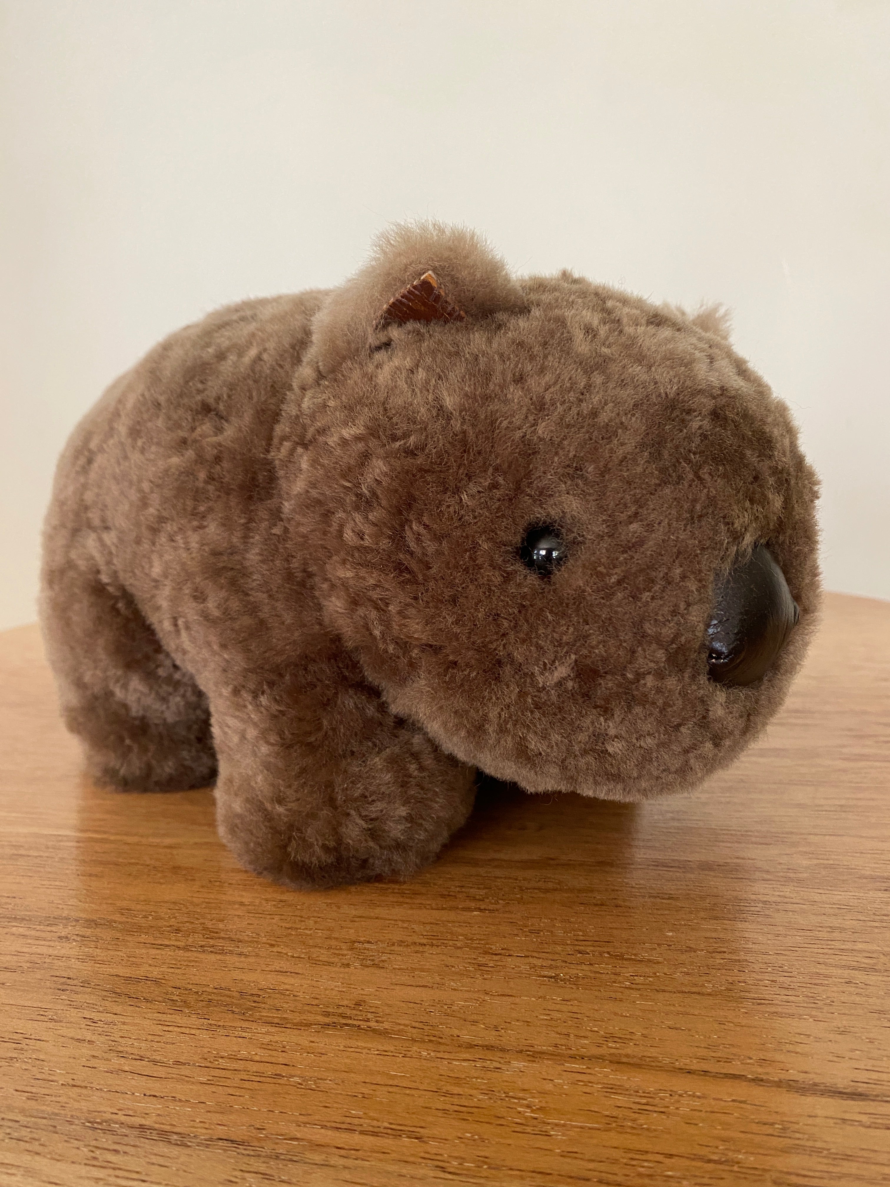 Winnie Wombat Sheepskin Soft Toy; shorn brown sheepskin with dark brown leather nose, plastic dark brown eyes and small brown leather inner ears and toes. Front view, angled to the left on a wooden table & against a pale wall.