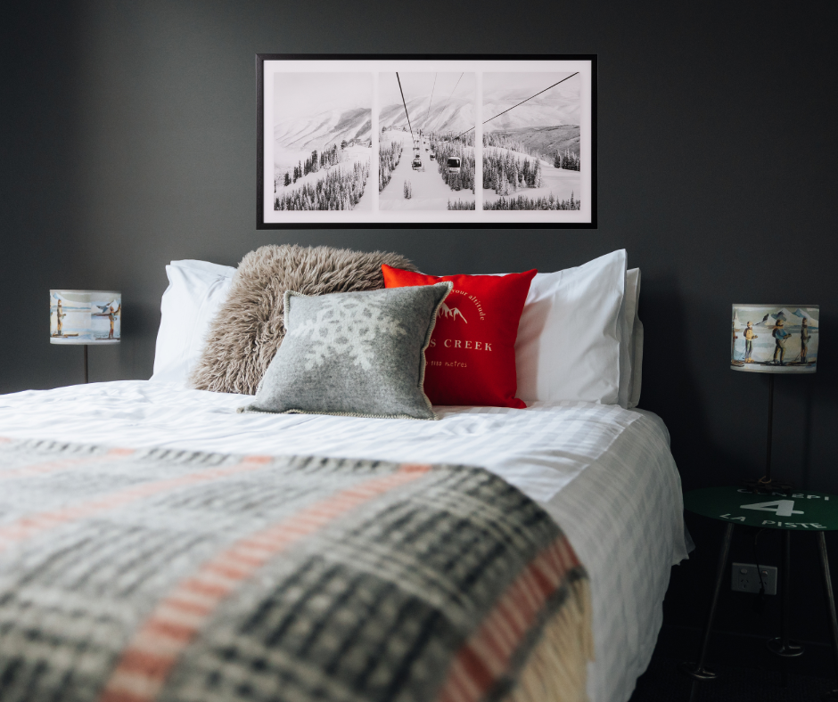 A stunning black and white triptych framed photograph of gondolas ascending a snow covered mountain in a black frame. A triptych is an image in 3 sections. Print show against a dark grey wall, above a large bed with white linen, grey, beige and red cushions and a black, grey, red and white blanket, green ski pole legged side tables and skier lamps.