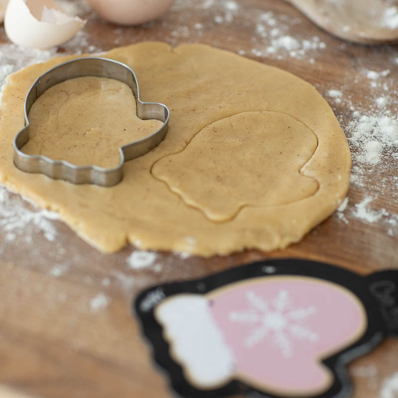 Image of mitten cookie cutter on wooden pastry board with pastry rolled out & the black cardboard cutter backing depicting a pink mitten with a snowflake resting off to one side