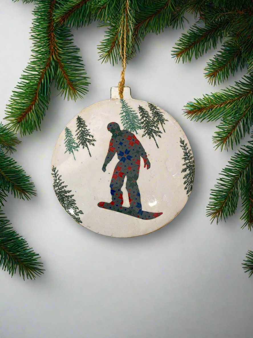 White metal hanging disc, with the image of a person snowboarding amongst trees. Front of disc.