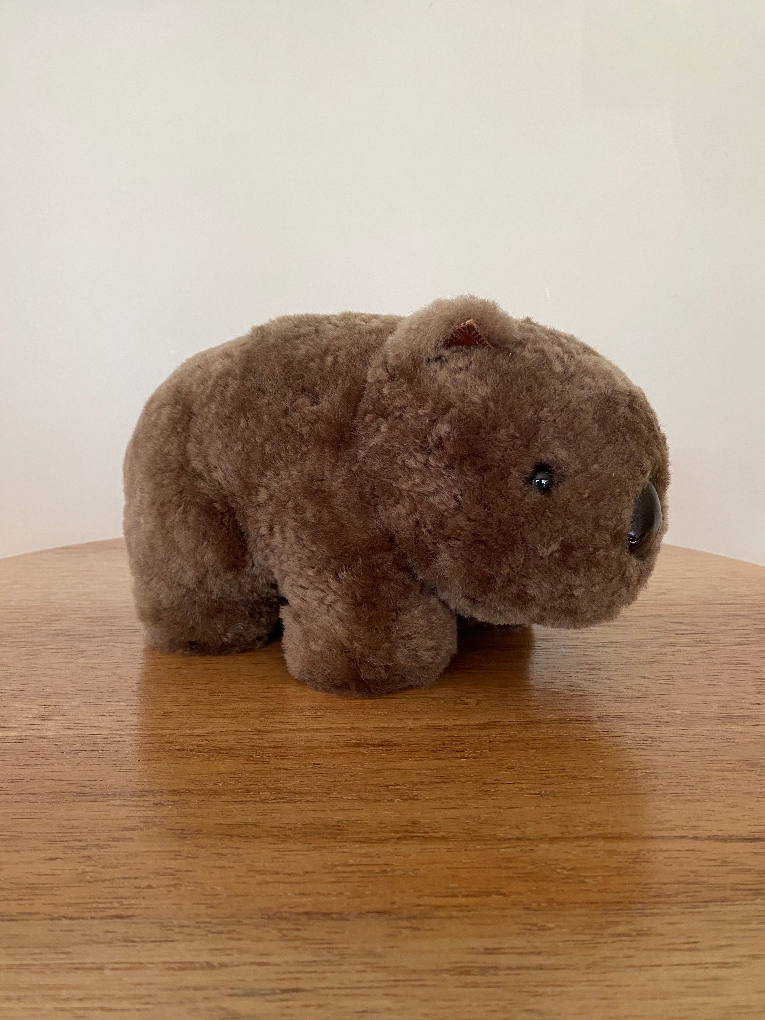 Winnie Wombat Sheepskin Soft Toy; shorn brown sheepskin with dark brown leather nose, plastic dark brown eyes and small brown leather inner ears and toes. Left hand side view, slightly angled to the left on a wooden table & against a pale wall.