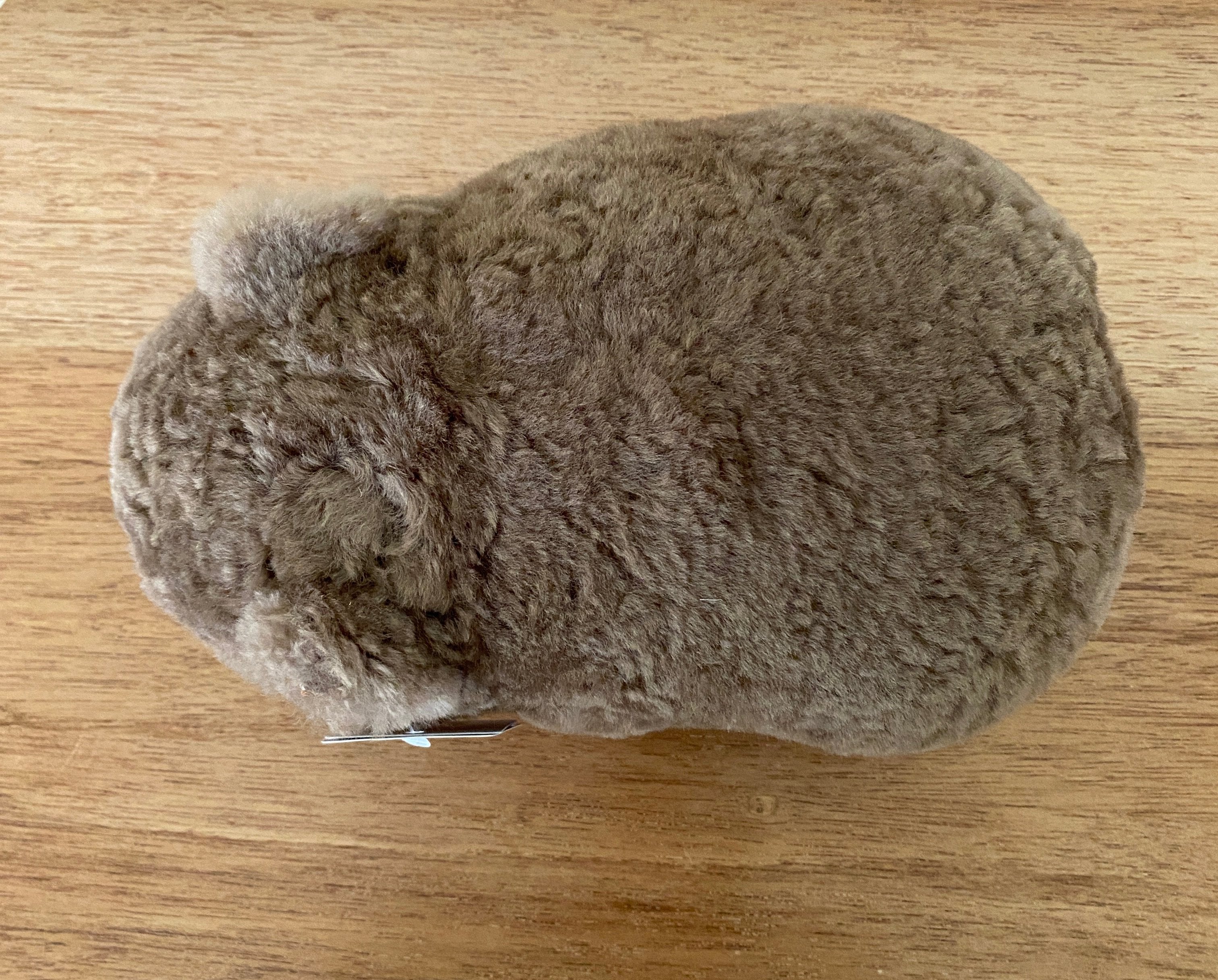 Winnie Wombat Sheepskin Soft Toy; shorn brown sheepskin with dark brown leather nose, plastic dark brown eyes and small brown leather inner ears and toes. Top down view, angled to the left on a wooden table.