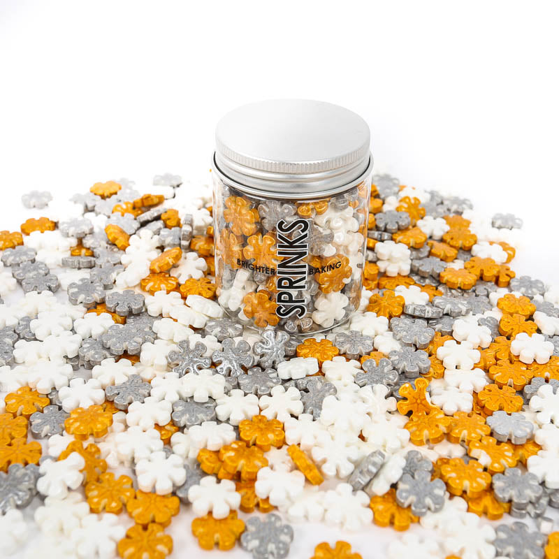 Image of a container of white, silver & gold snowflake shaped sprinkles with extra sprinkles scattered around the container. Sprinkles for cake or cookie decorating viewed from the front with brand label visible 
