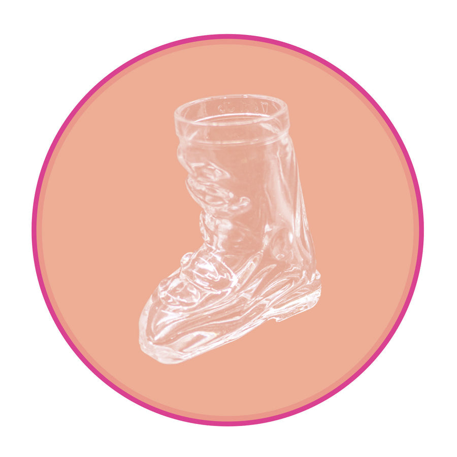 Photo of a single Après Allstar boot shot glass. The shot glass is made of clear glass. Image is set against a peachy pink backdrop.