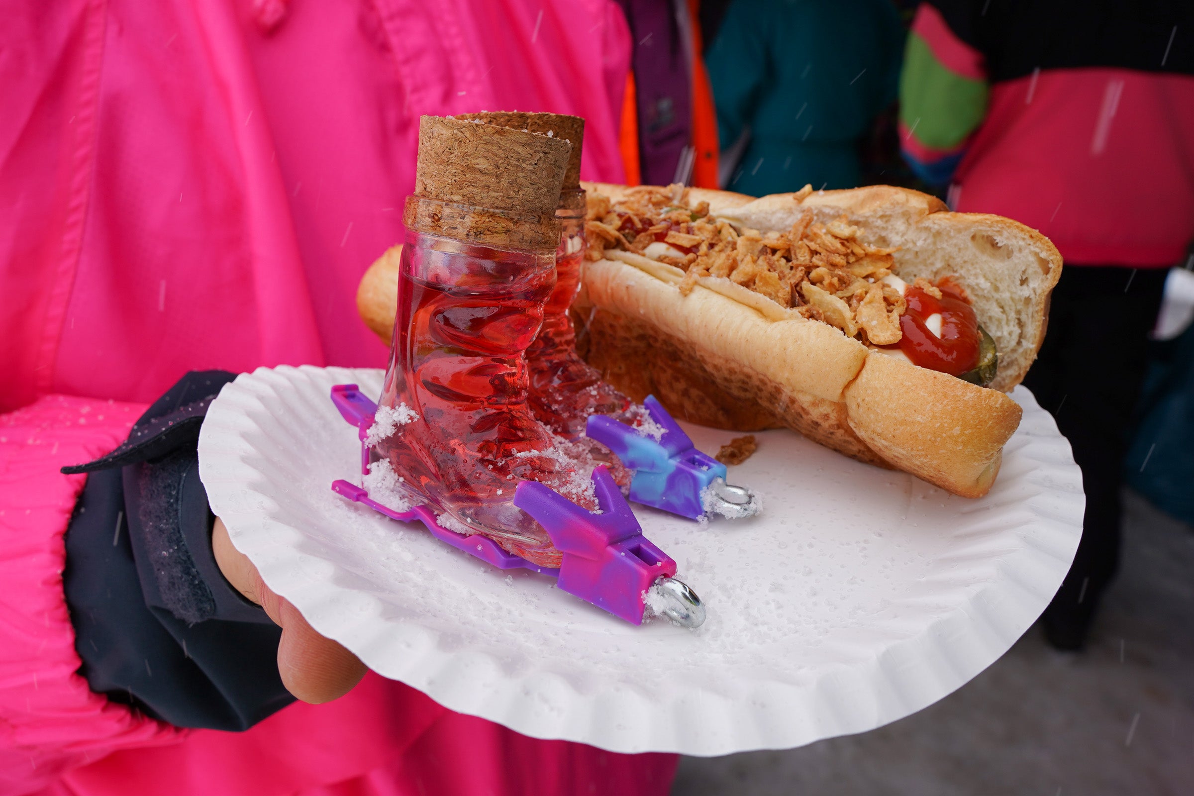 Photo of 2 Apr√®s Allstar ski boot shot glasses, coloured binding & cork, filled with liquid & sitting on a paper plate with a hotdog being held by someone in a pink and green outfit.