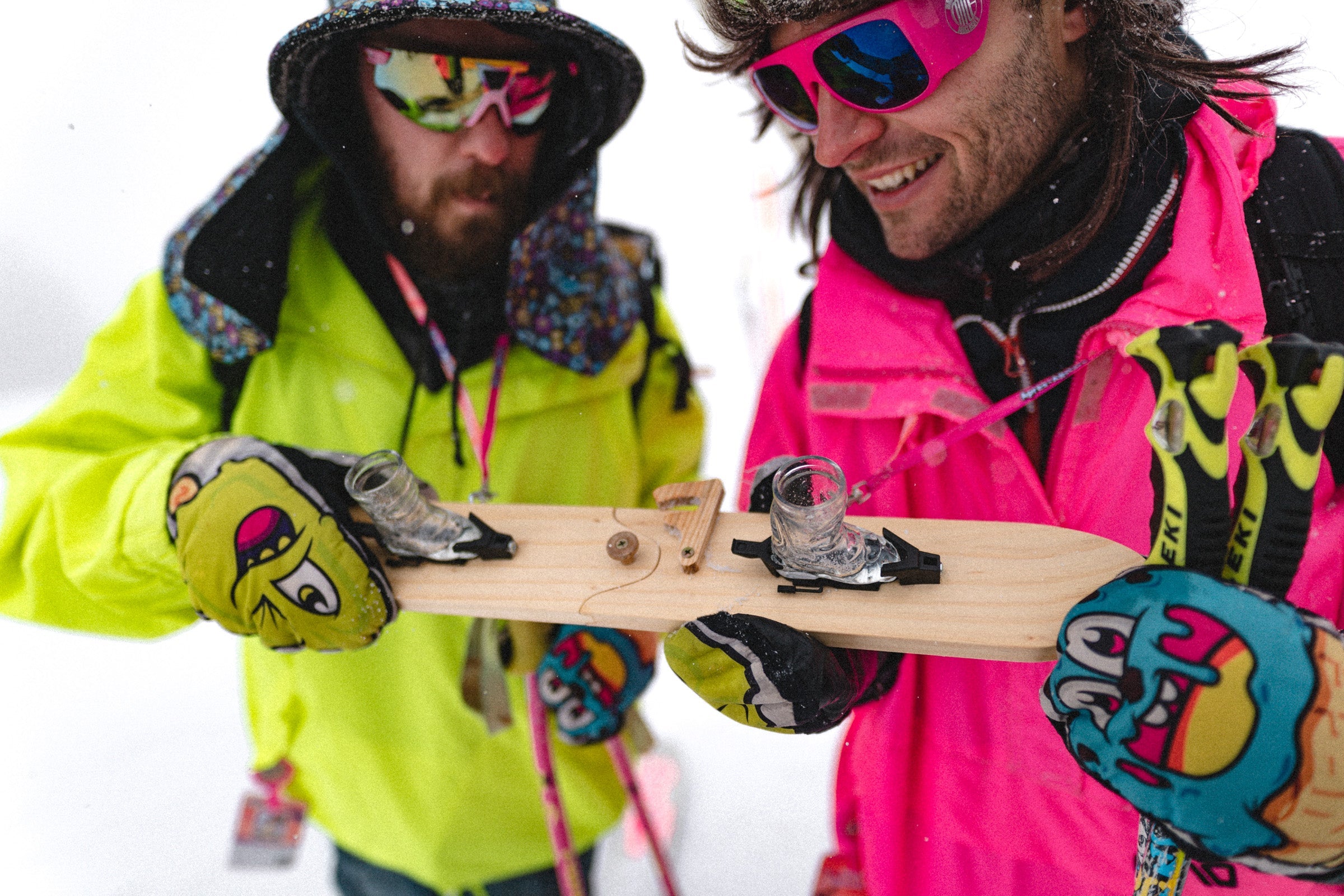 Photo of of Michi & Tobi, the Apr√®s Allstar founders, in a yellow & pink jackets, colourful hats and sunglasses with part of an Apres Allstar SwigSki3000 fitted with the bindings and ski boot shot glasses held between them.