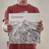 A person wearing a red t-shirt holds up a copy of The Many Behinds The Maps book featuring a detailed black and white sketch of mountains, with the text 'THE MAN BEHIND THE MAPS' visible at the top.