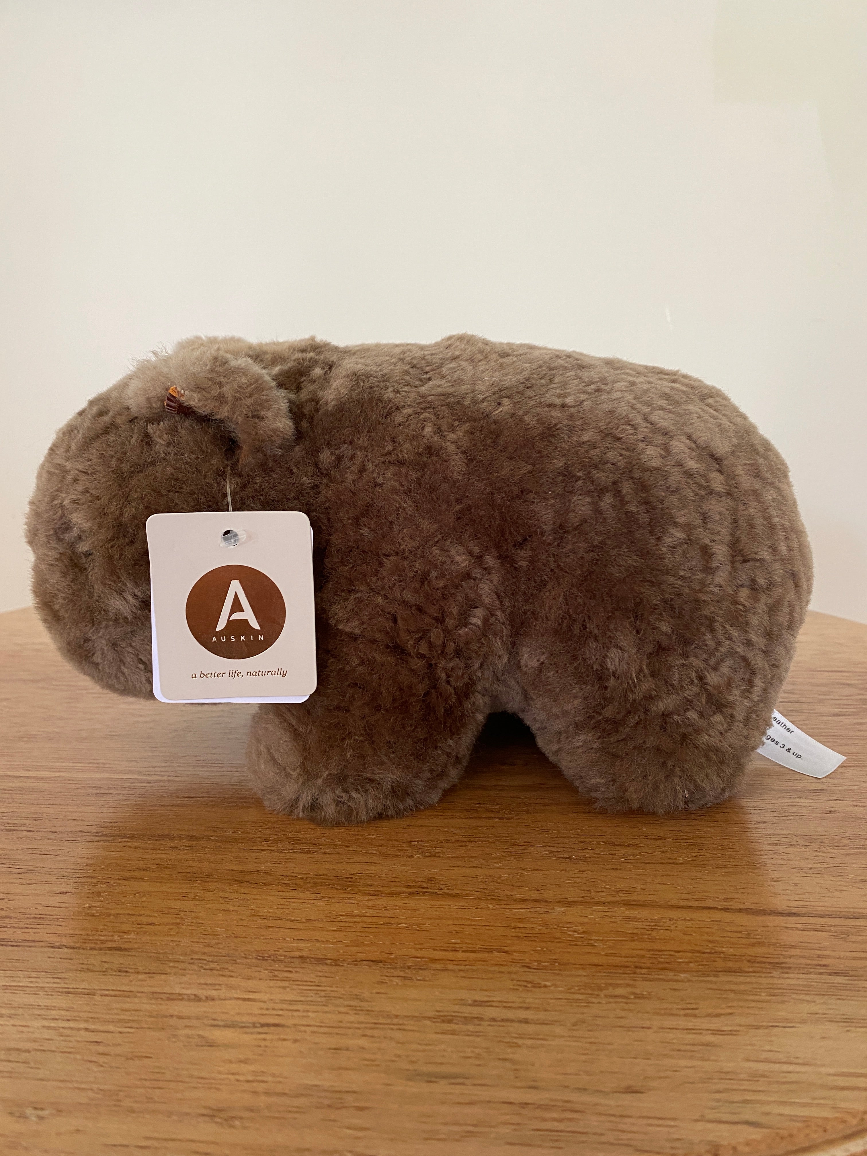 Winnie Wombat Sheepskin Soft Toy; shorn brown sheepskin with dark brown leather nose, plastic dark brown eyes and small brown leather inner ears and toes. Right hand side view, angled to the left on a wooden table & against a pale wall. Brand sales label attached to ear.