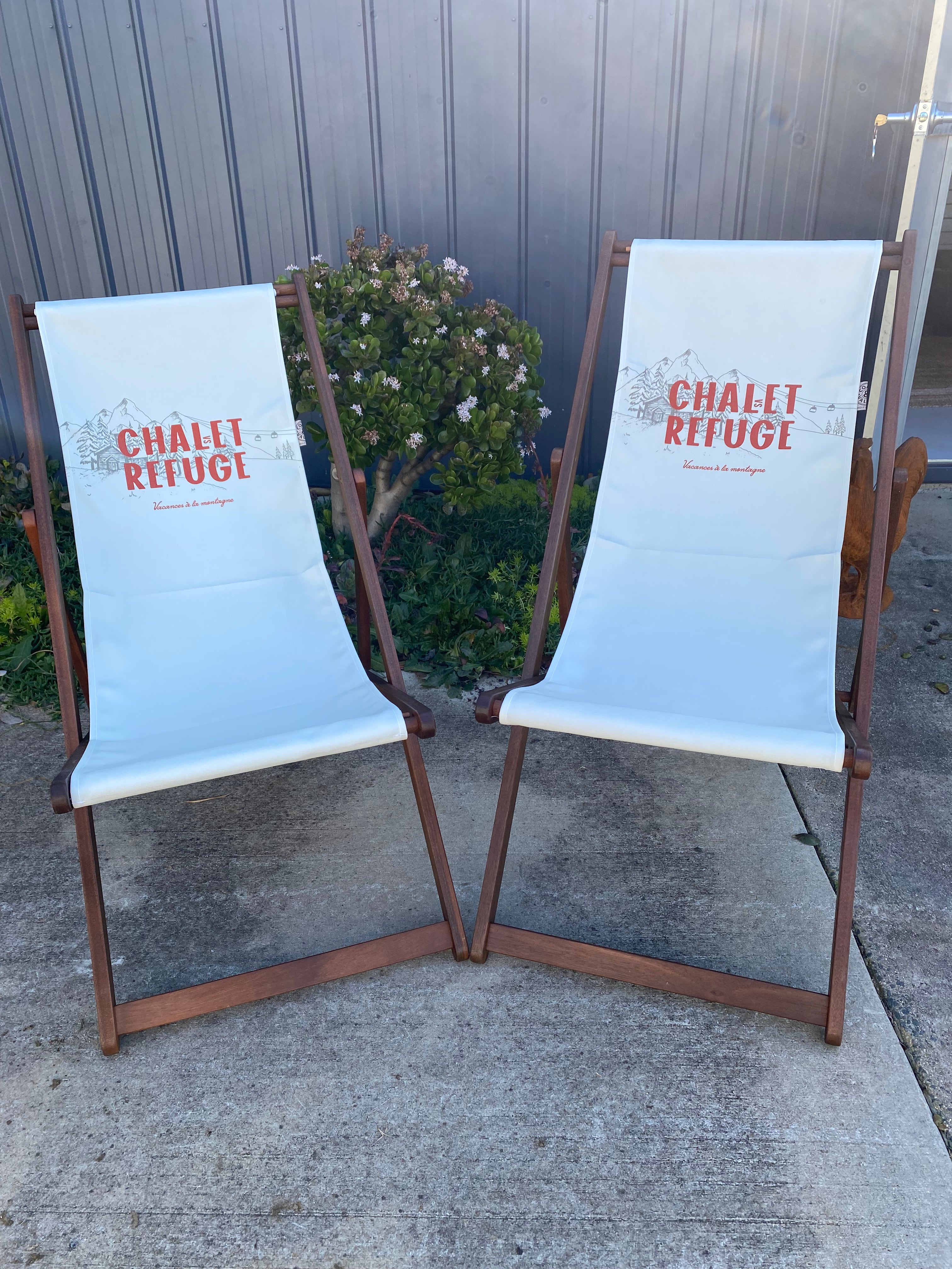 Pair of Coast & Valley Deck Chairs: Chalet vs Refuge design showing words in red with a grey grey chalet surrounded by trees on a plain cream background.2 chairs facing forward. On a concrete floor against a blue steel shed and door with garden bed in the background.