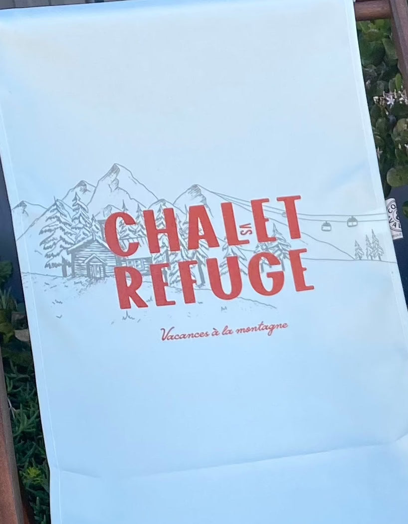Coast & Valley Deck Chair Sling: Chalet vs Refuge design showing words in red with a grey grey chalet surrounded by trees on a plain cream background. Sling close up. On a concrete floor against a blue steel shed and door with garden bed in the background.
