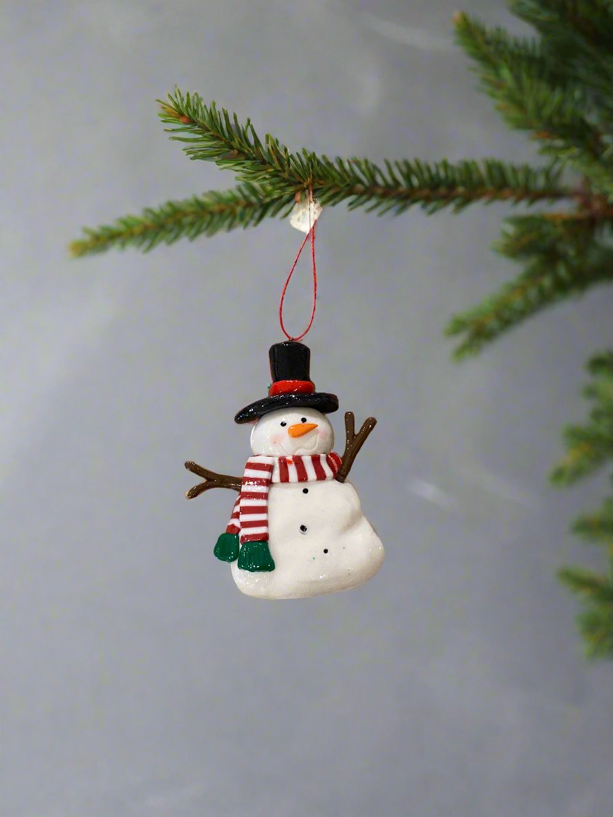 small white snowman ornament, wearing a black hat with a red hat band, brown sticks for arms, a red &amp; white striped scarf with a green fringe, and with black dots down his chest and an orange nose, hanging from a tree branch