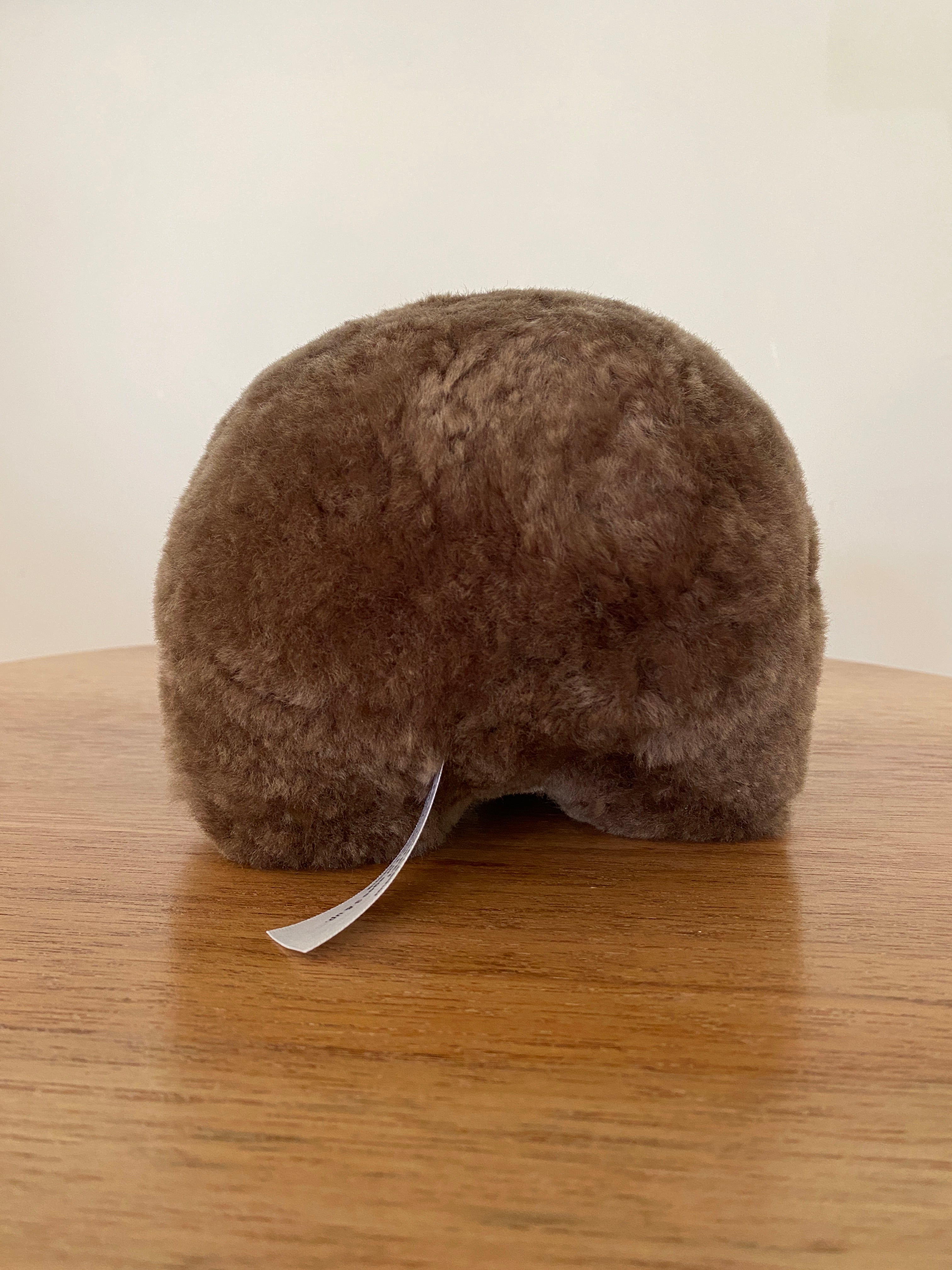 Winnie Wombat Sheepskin Soft Toy; shorn brown sheepskin with dark brown leather nose, plastic dark brown eyes and small brown leather inner ears and toes. Rear view, on a wooden table & against a pale wall.