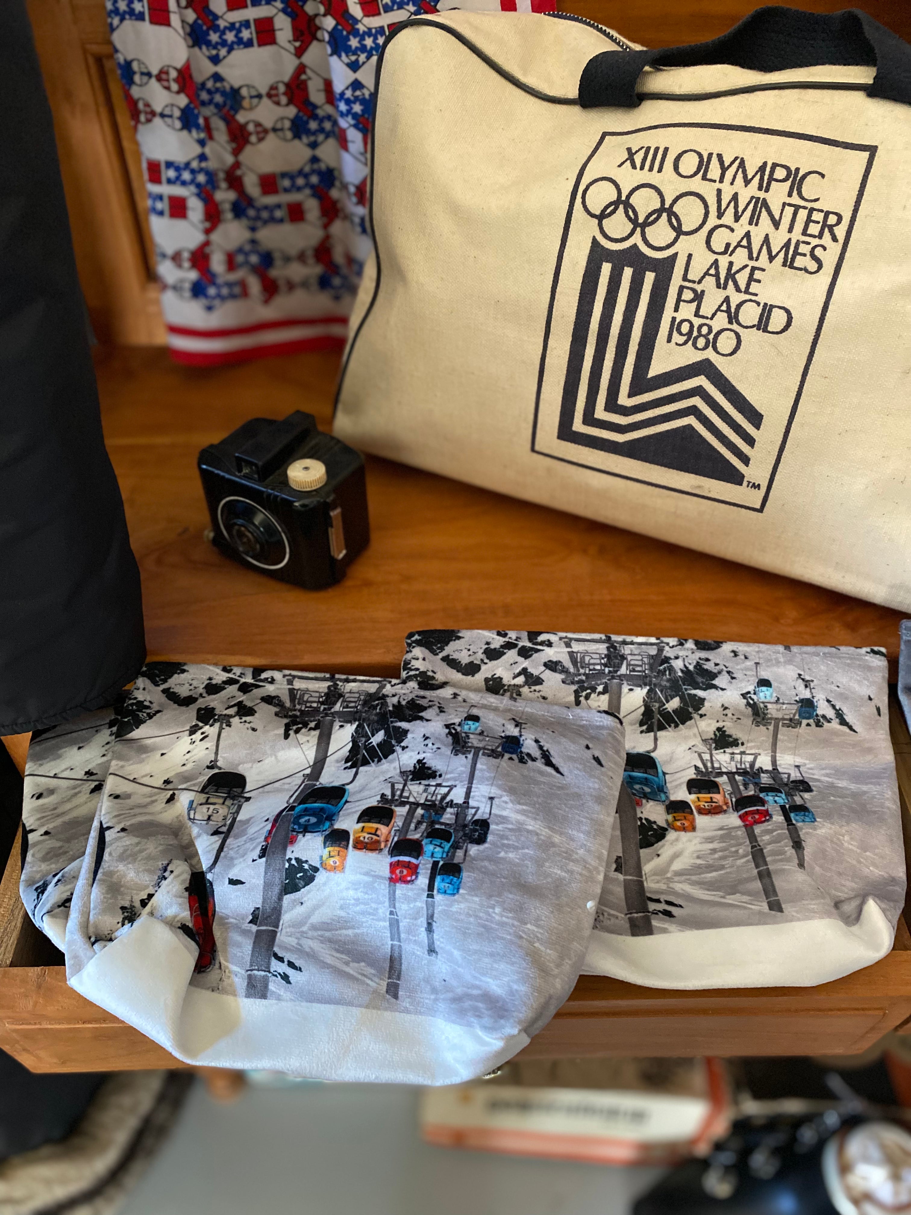 Photo of coloured gondola against the backdrop of a ski slope resting on a wooden table with a vintage Bakerlike Kodak Brownie Camera and a 1980 Lake Placid overnight bag in the background