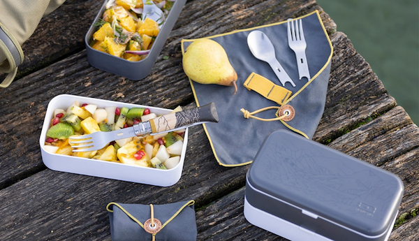 outdoor lunch box filled with food and the cutlery handle and knife/spoon/fork kit and napkin on wooden picnic table 