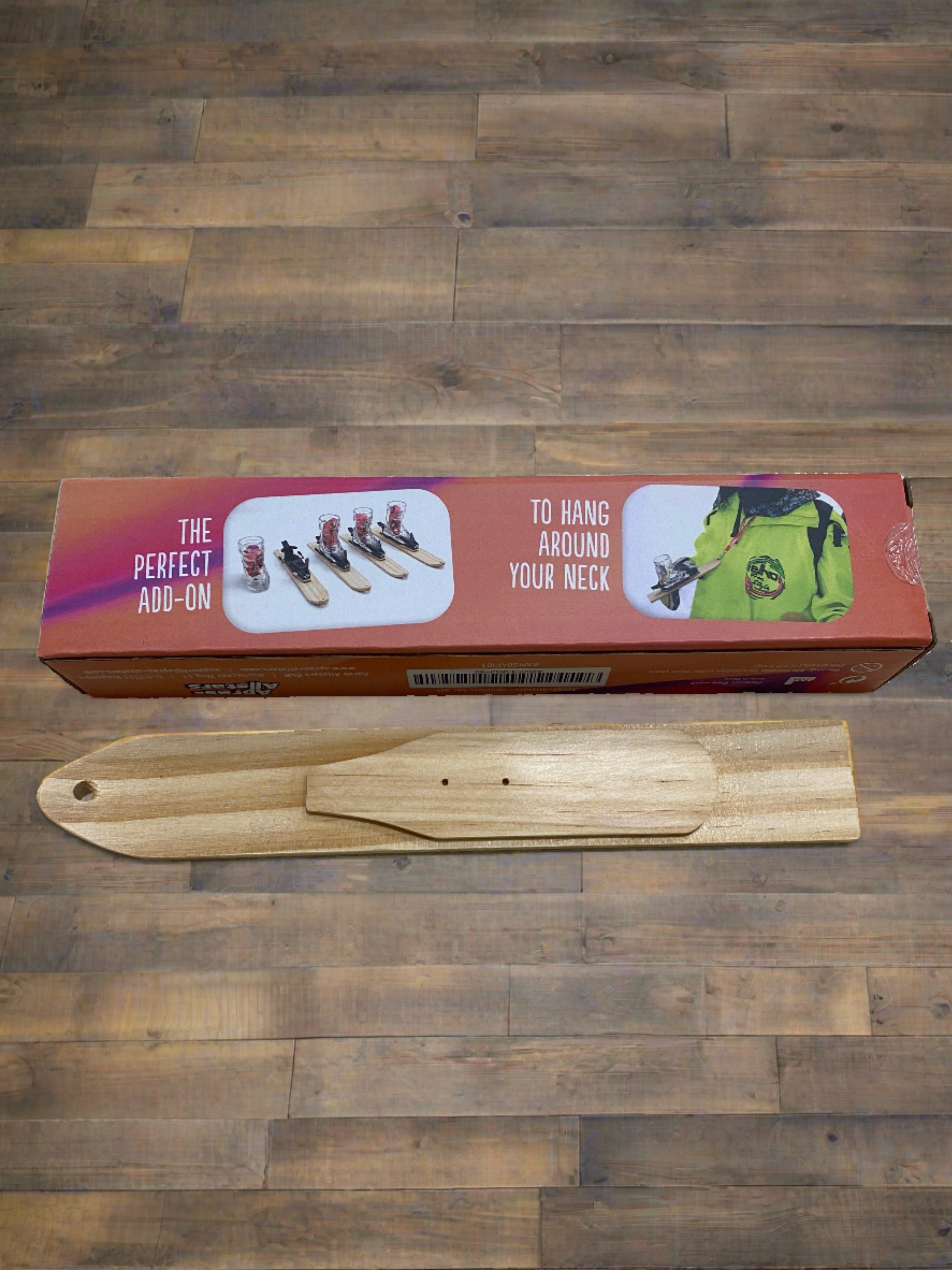 Photo of a wooden tray in the shape of small ski & the box it comes in. The ski tray has a hole drilled into the tip of the ski and a small raised mounting platform for the Apres Allstar boot shot glass and bindings. Image is set against a wooden backdrop