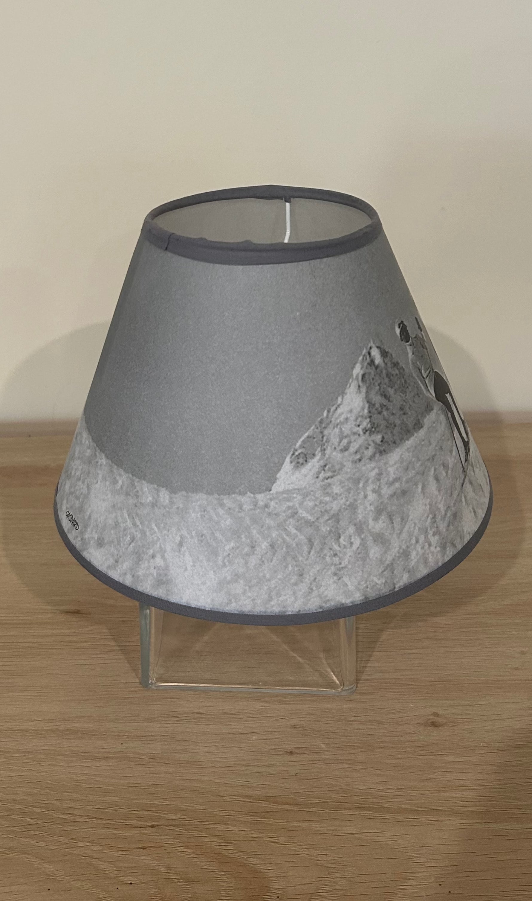 This lamp shade is a vintage black &amp; white photo two men and a woman standing on skis supporting themselves with vintage single long poles, against a backdrop of snow covered mountains.. On a wood table top. Side view