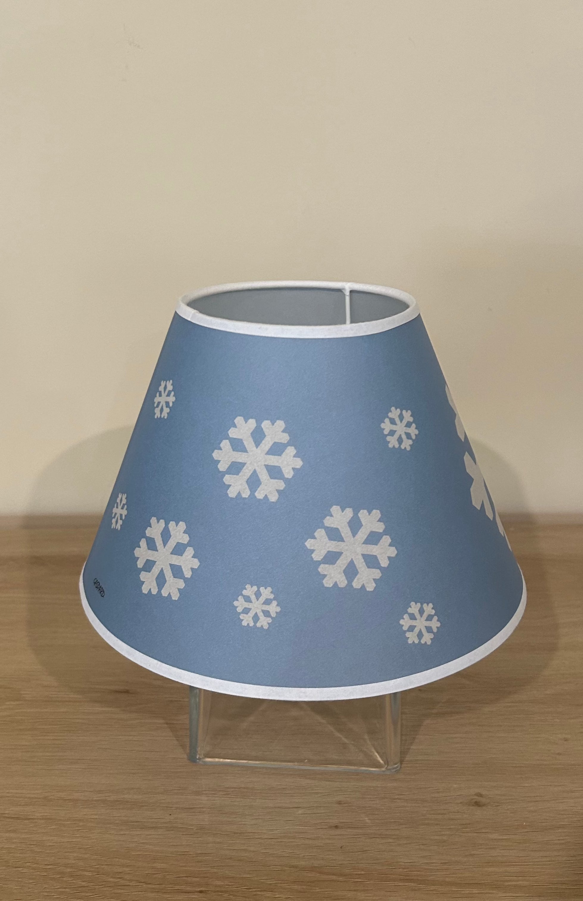 La Maison De Gaspard lamp shade with a snowflake printed canvas lamp shade in blue and white. On wooden table, resting on glass cube. Different angle