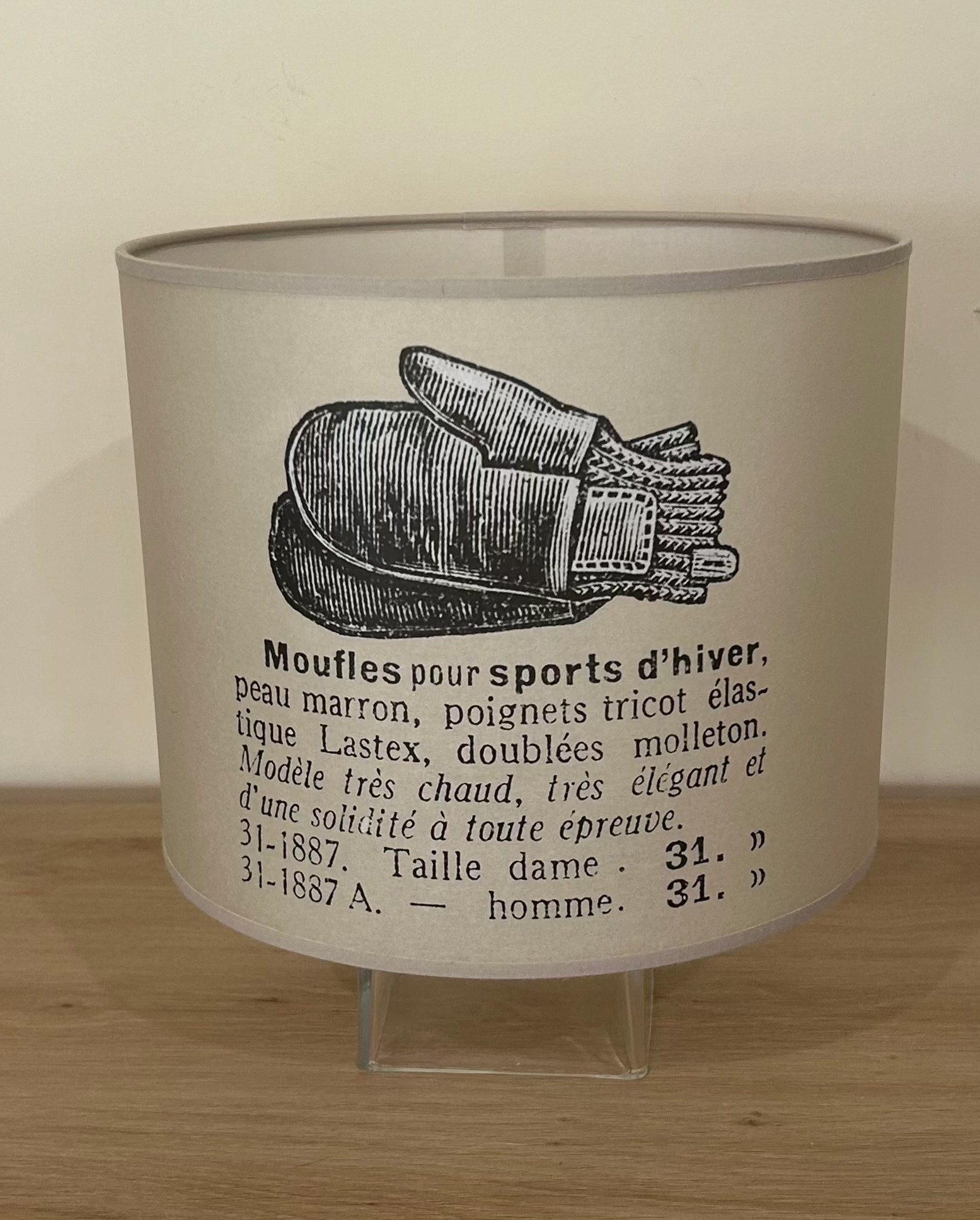 La Maison De Gaspard canvas printed beige lamp shade with a vintage black &amp; white drawing of a pair of woollen mittens and description in french of the mittens. On wooden table, on top of a glass cube