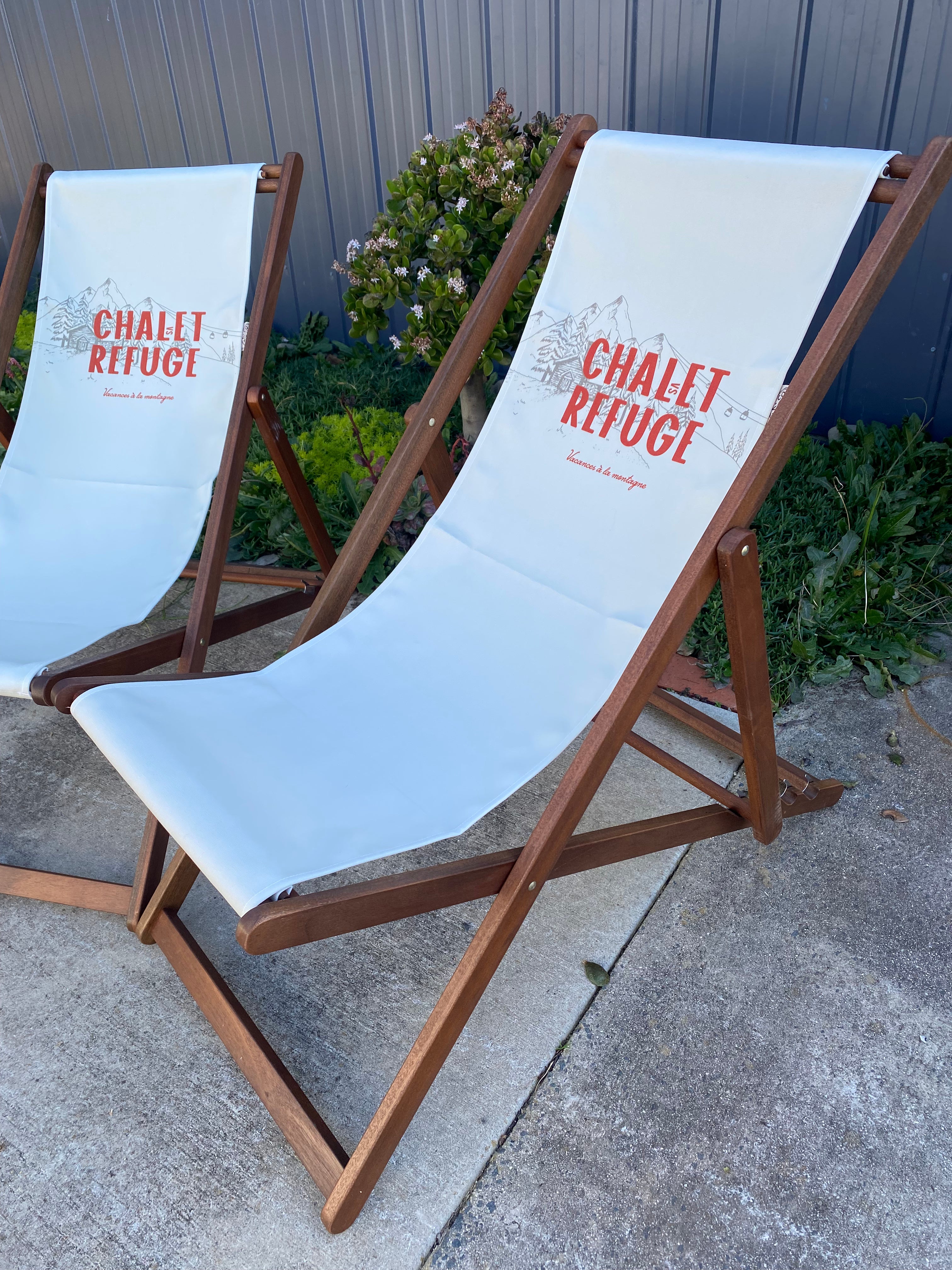 Pair of Coast & Valley Deck Chairs: Chalet vs Refuge design showing words in red with a grey grey chalet surrounded by trees on a plain cream background. 2 chairs side on view. On a concrete floor against a blue steel shed and door with garden bed in the background.