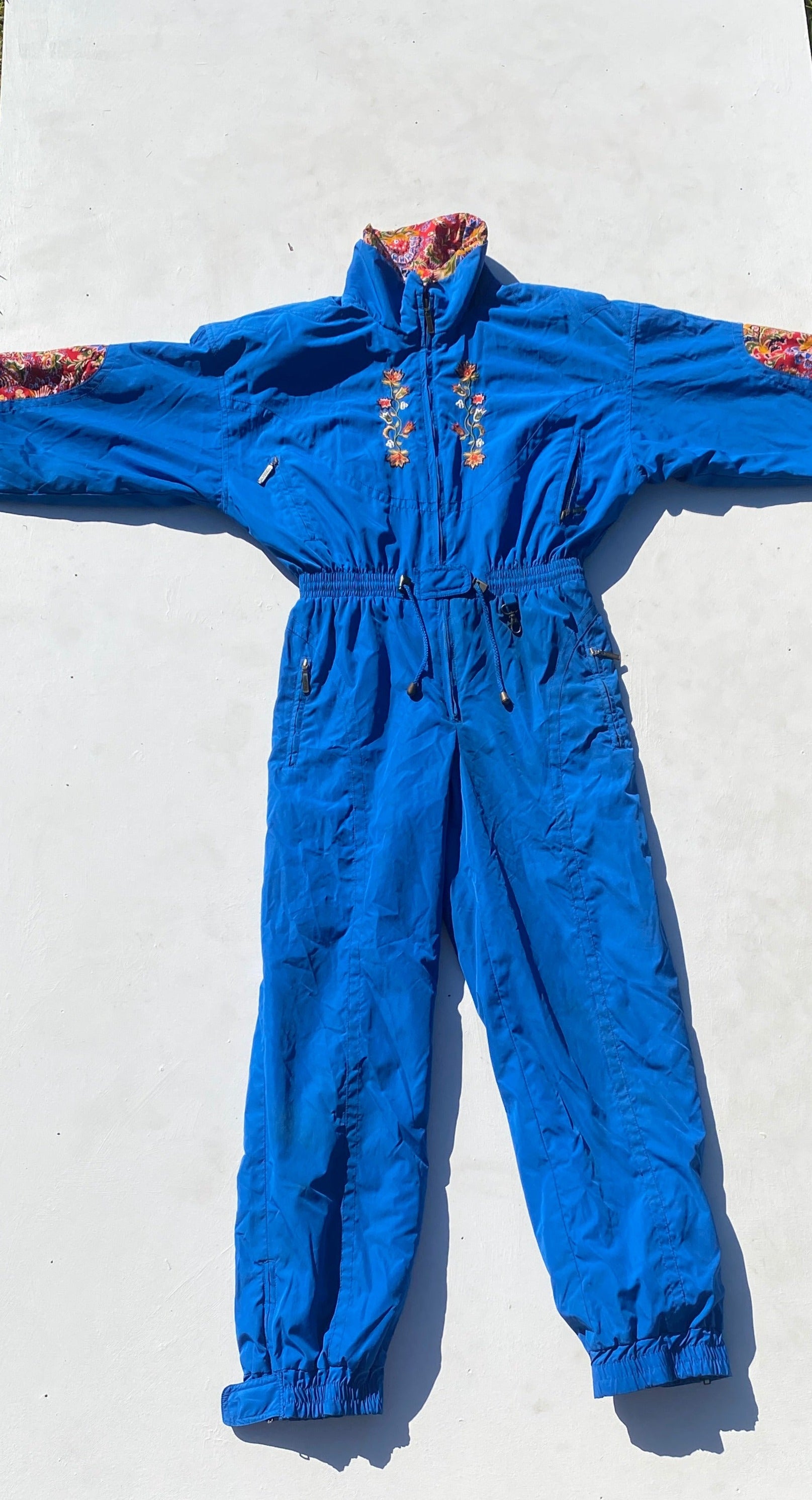 Powderhorn royal blue embroidered one piece ski suit with red, yellow, green and blue patterned floral fabric patches, with drawstring waist. Front view flat lay