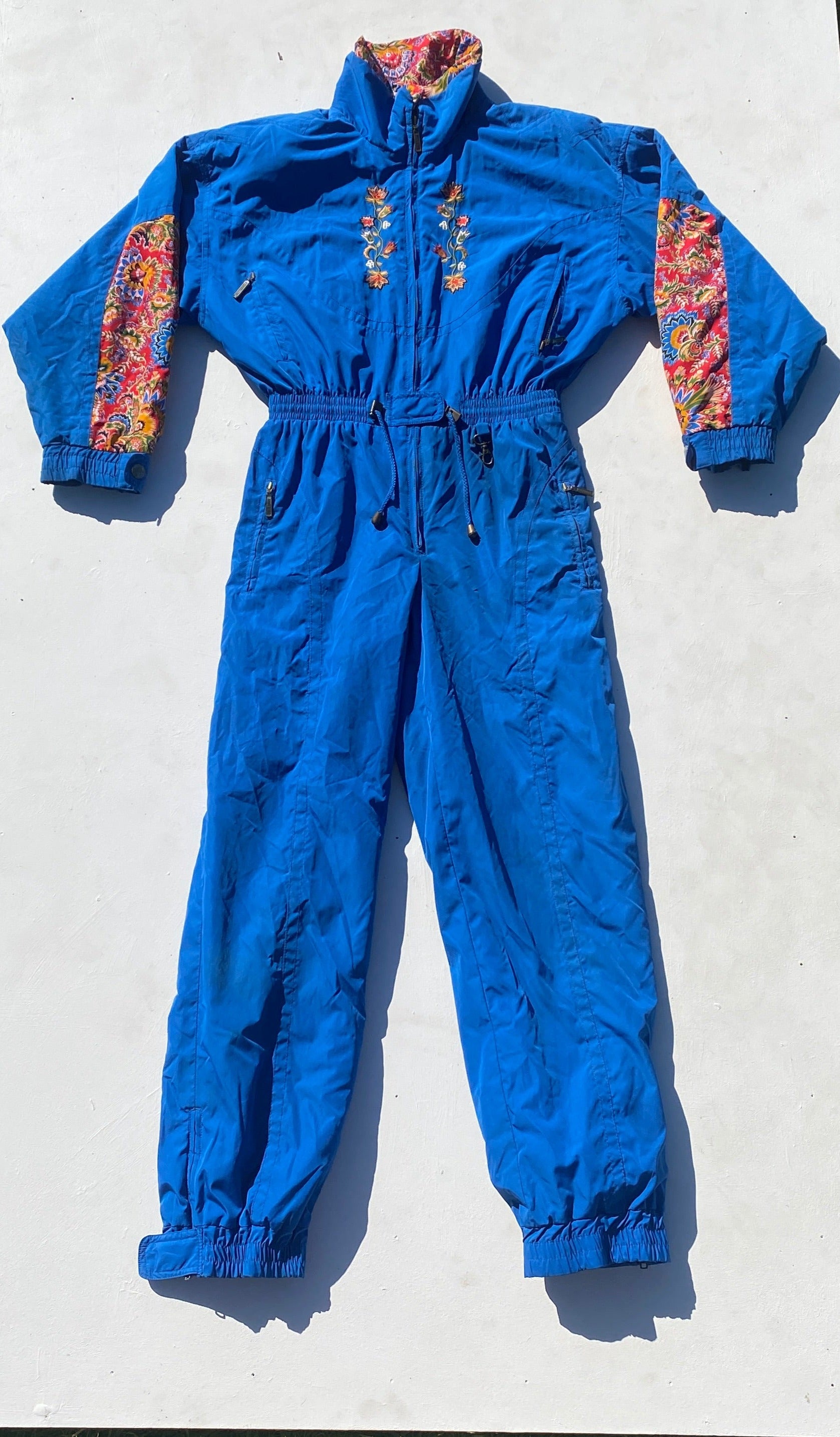 Powderhorn royal blue embroidered one piece ski suit with red, yellow, green and blue patterned floral fabric patches, with drawstring waist. Front view flat lay arms bent