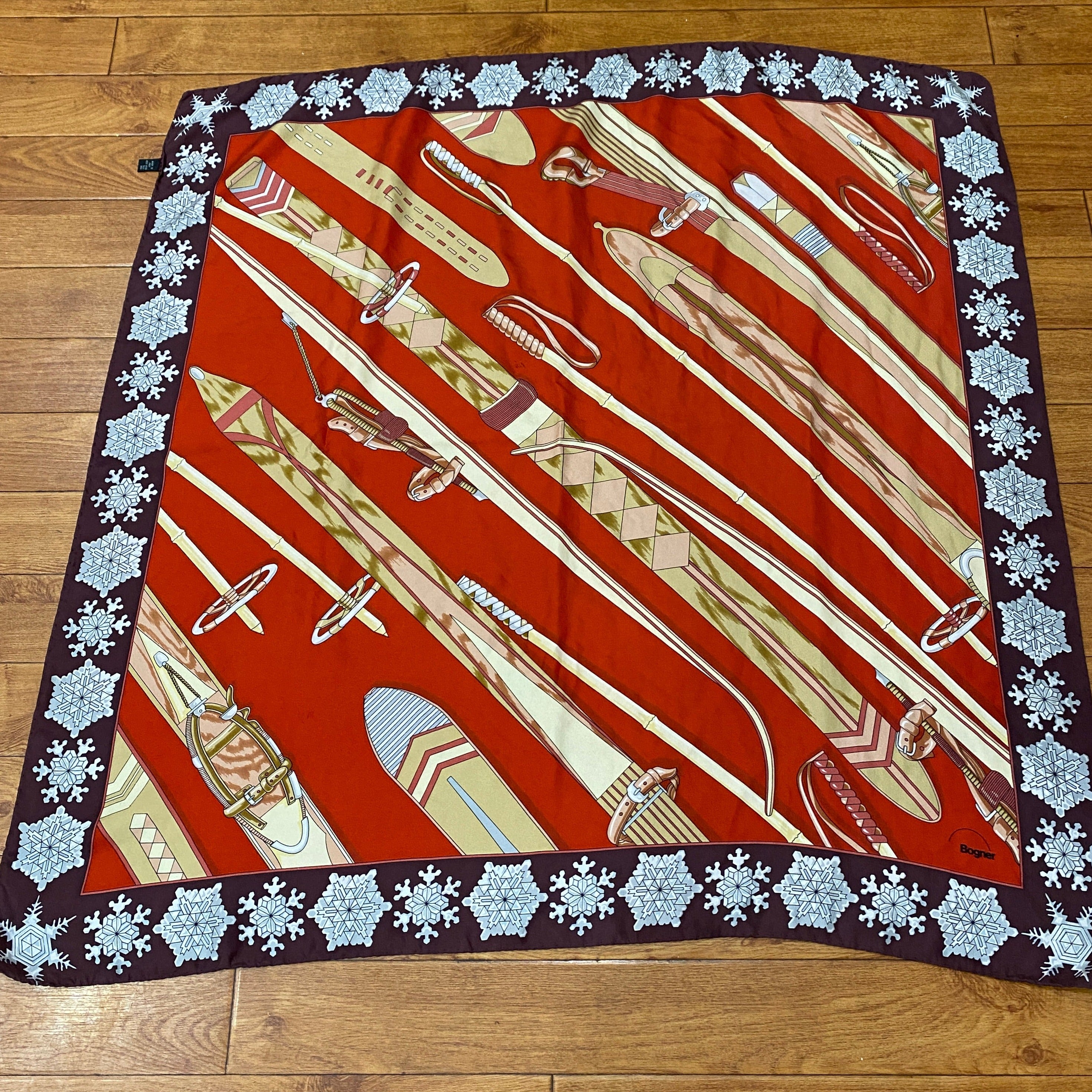 Bogner Silk Vintage Ski Scarf in red, brown white and beige. Snowflakes border the scarf, with the central square illustrating wooden skis and poles. Shown against a wooden background.