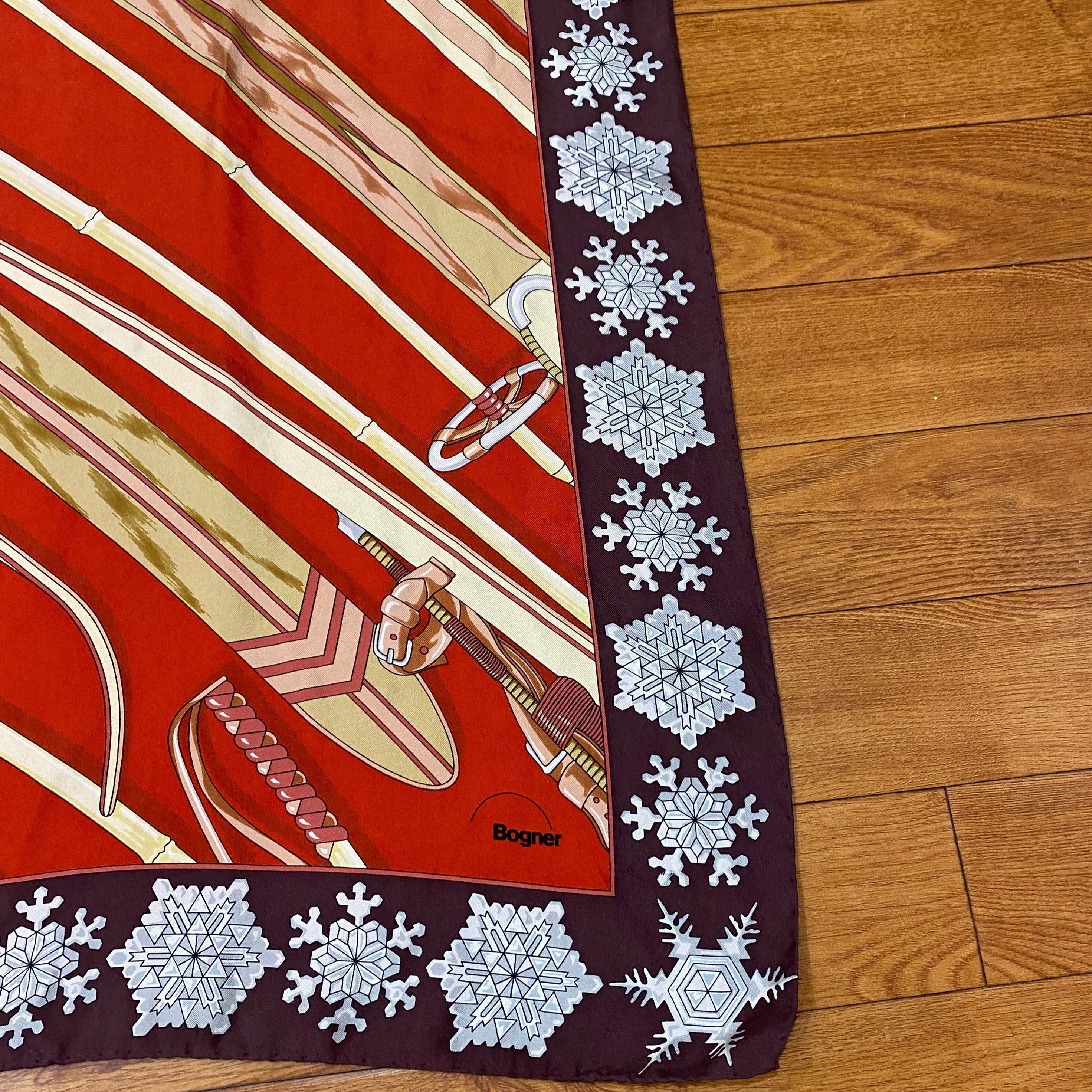 Bogner Silk Vintage Ski Scarf in red, brown white and beige. Snowflakes border the scarf, with the central square illustrating wooden skis and poles. Lower Right corner of scarf showing Bogner brand.Shown against wooden background