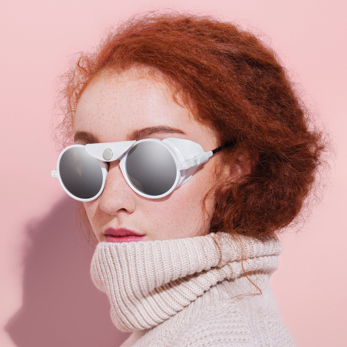 Image of a red haired woman wearing a pair of white IZIPIZI Gun Glacier framed folded sunglasses and a pale pink roll necked knitted top. The glasses have a leatherette flap across the nose bridge of the glasses, attached with a silver press stud, & across the arms of the glasses to block the sun's glare. Set against a pink background.