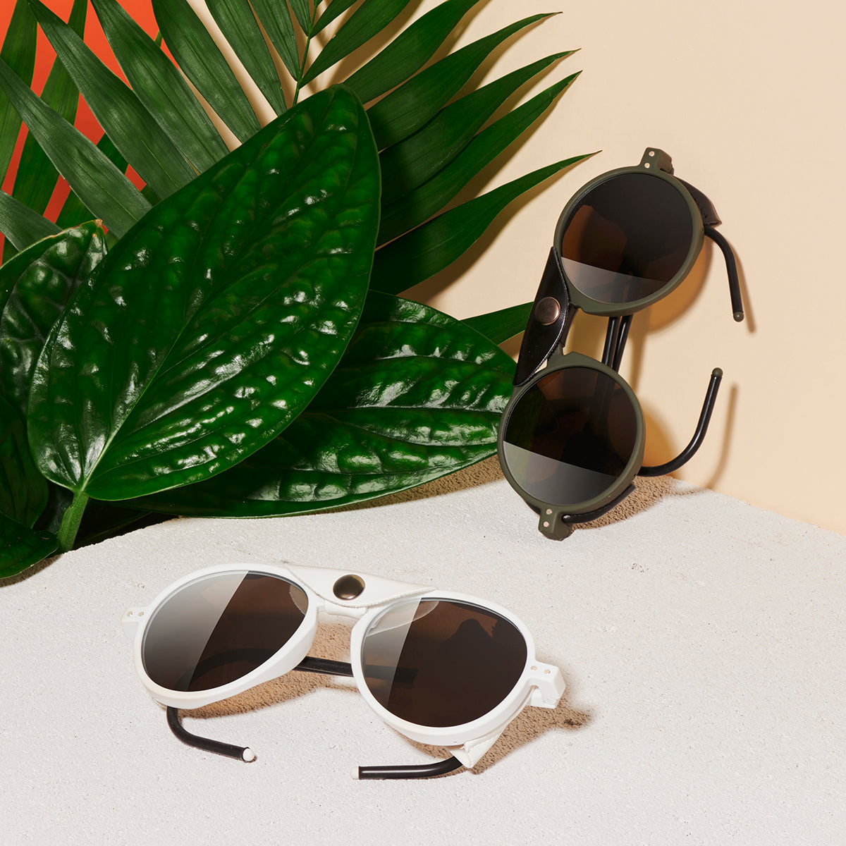 Image of 2 pairs of sunglasses. 1 white framed folded sunglasses and 1 dark brown pair, with with black arms & a leatherette flap across the nose bridge of the glasses & attached with a silver press stud. Set on a white ledge against a wall with green foliage to the left hand side.
