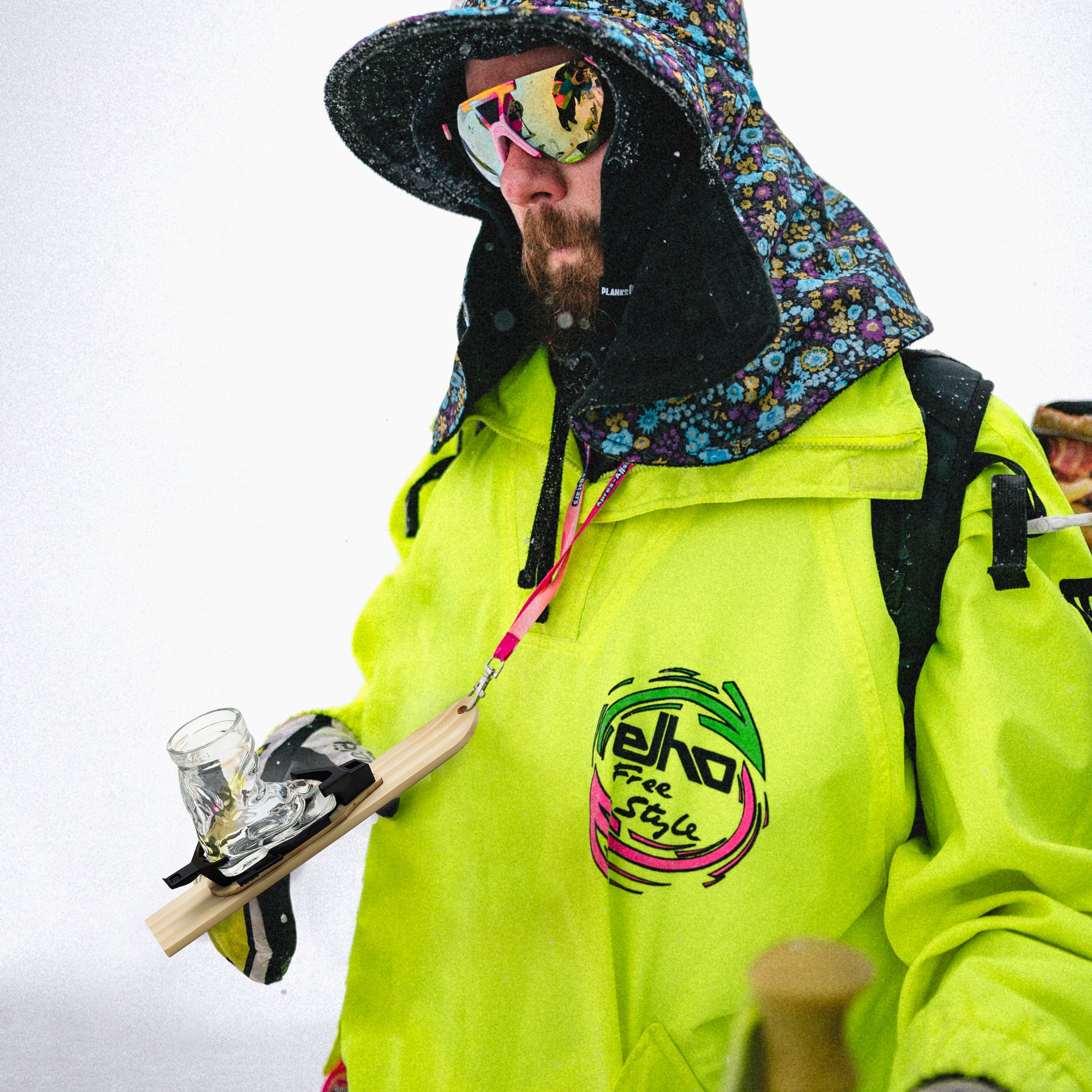 Photo of Michi, 1 of the Apres Allstar founders, in a yellow jacket, colourful hat and sunglasses wearing his mini-ski on a Apr√®s Allstar lanyard around his neck in the snow. The mini-ski has been fitted out with the Apr√®s Allstar bindings boot shot glass & lanyard.