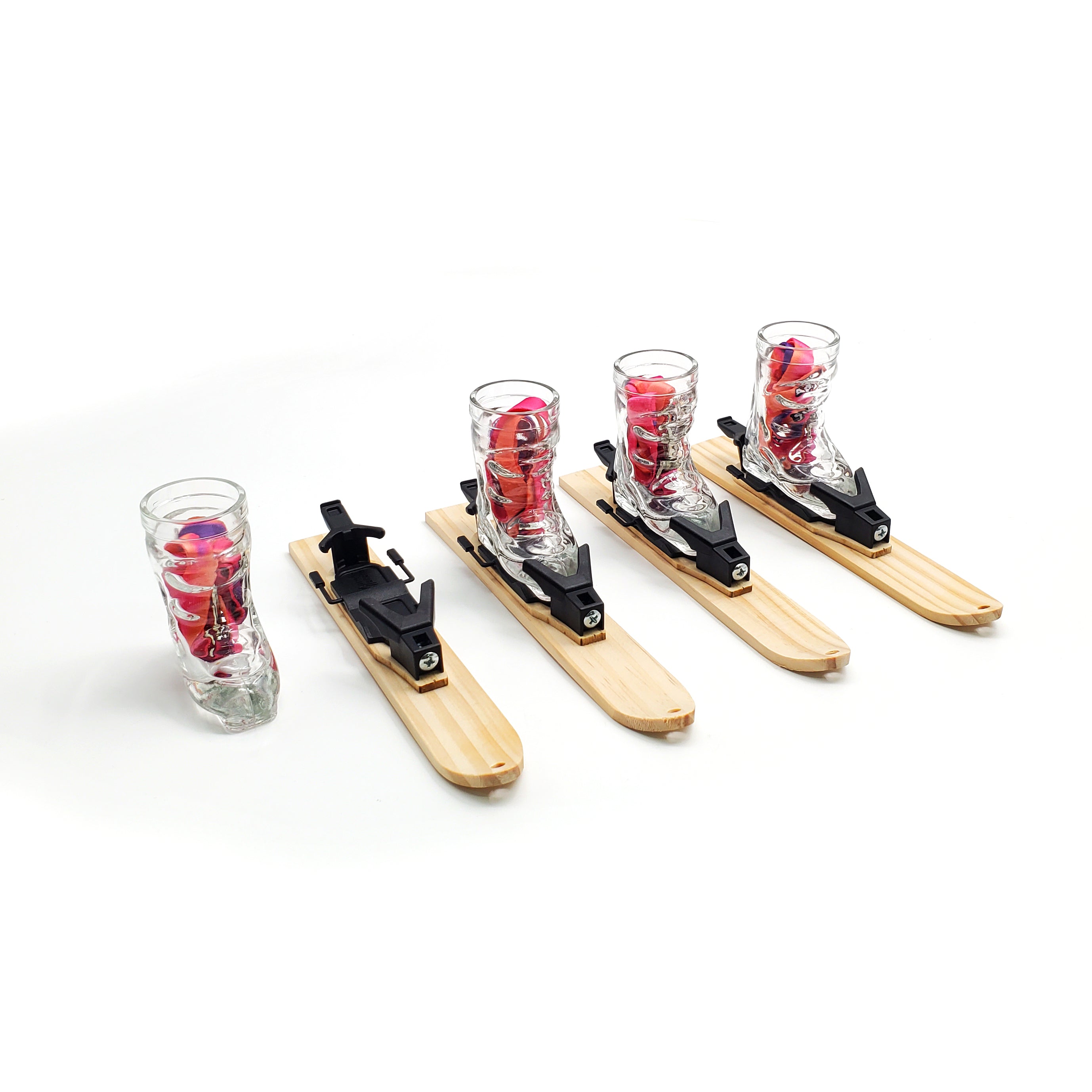 Photo of 4 wooden trays in the shape of small skis. Each ski tray has a hole drilled into the tip of the ski and a small raised mounting platform for the Apres Allstar boot shot glass and bindings that are shown mounted on the mini skis. A red lanyard is sitting inside the glass. Image is set against a white backdrop