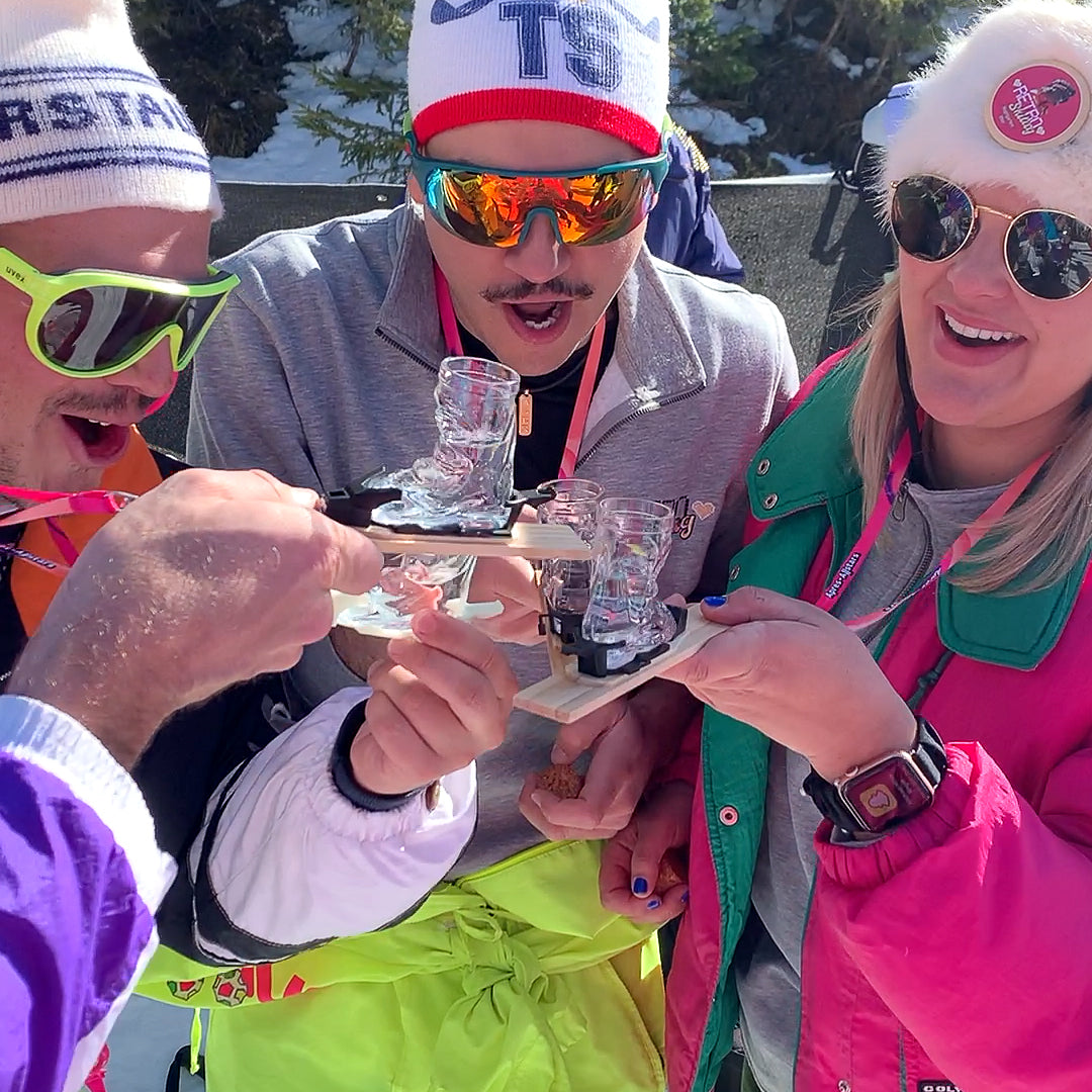 Photo of 2 men with moustaches and a blond haired lady in colourful ski gear and sunglasses, wearing the Apr√®s Allstar mini-ski on a lanyard around their necks in the snow. The mini-skis have been fitted out with the Apr√®s Allstar bindings boot shot glass & lanyard.
