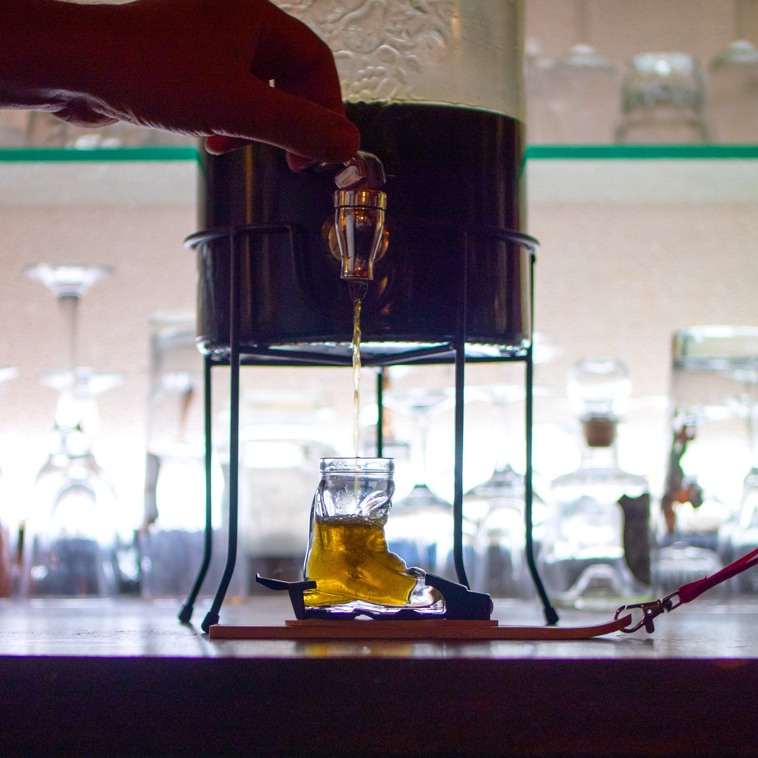 Photo of the mini-ski, fitted with the ski boot shot glass and bindings, underneath a glass drinks dispenser. A hand touches the lever pour the liquid from the dispenser into the shot glass. Against a backdrop of shelves of bar glassware