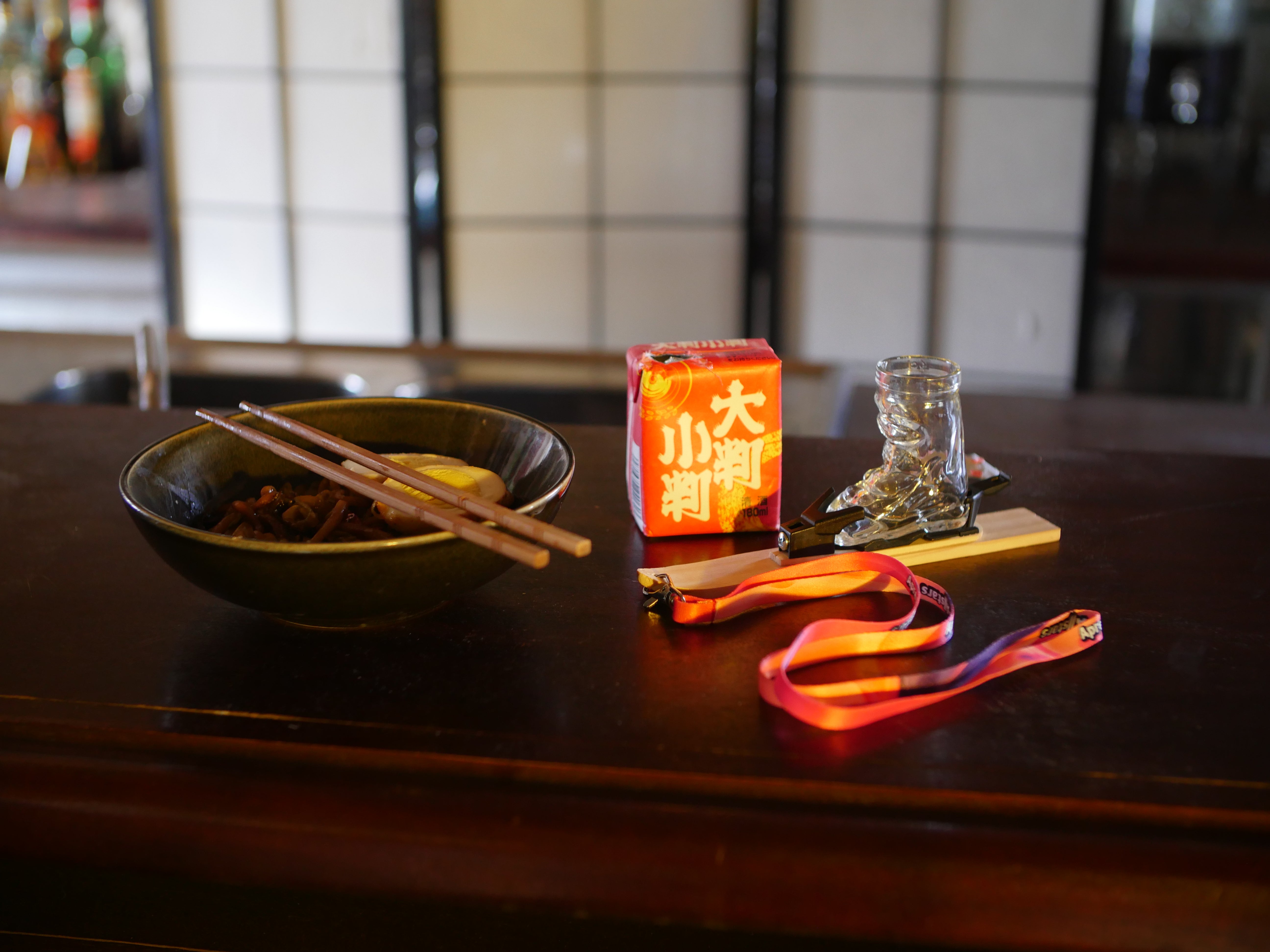 Photo of the mini-ski, fitted with the ski boot shot glass and bindings, on a wooden table in front of a Japanese style white screen/room divider. The table is set with a bowl of food, a pair of chopsticks and a small red box with asian symbols.