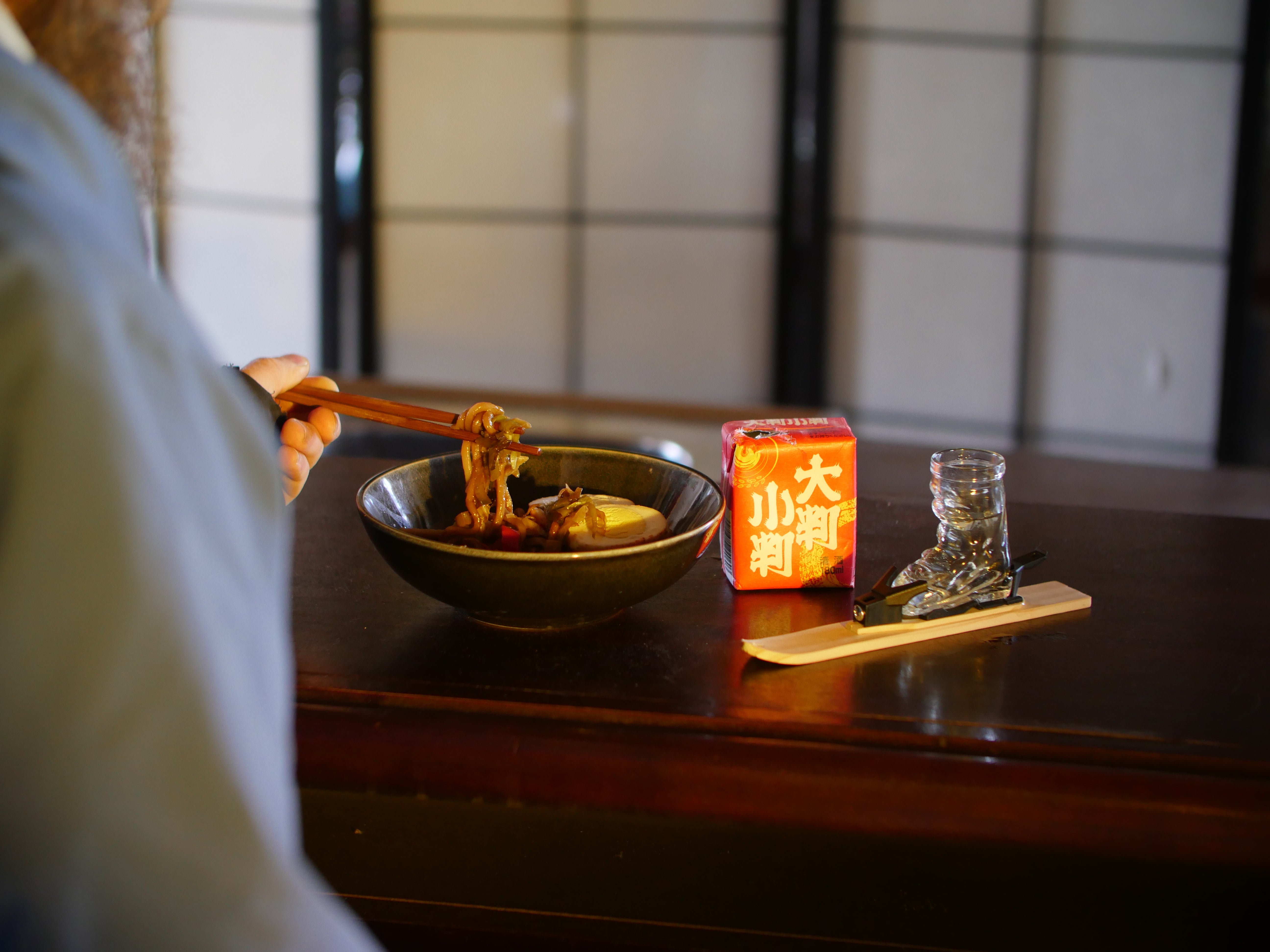 Photo of the mini-ski, fitted with the ski boot shot glass and bindings, on a wooden table in front of a Japanese style white screen/room divider. The table is set with a bowl of food, a pair of chopsticks and a small red box with asian symbols. A person in white is lifting the chopsticks filled with food.