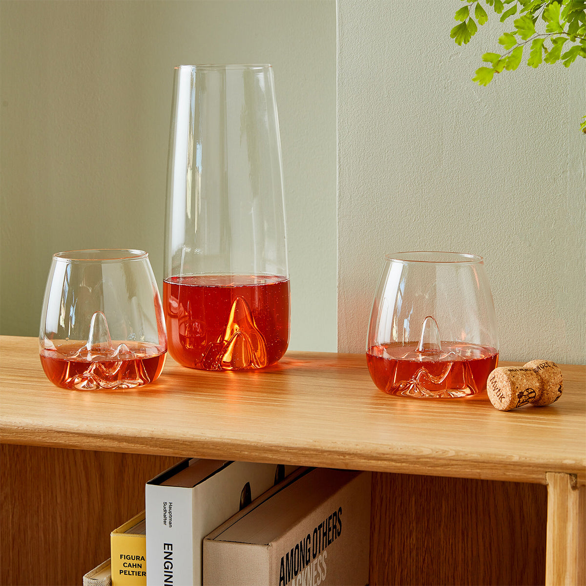 Image of a glass carafe and a pair of glass tumblers filled with an orange liquid sitting on a timber bookshelf against a light olive green wall. The carafe & glasses are indented with the shape of a mountain peak indented into their base.