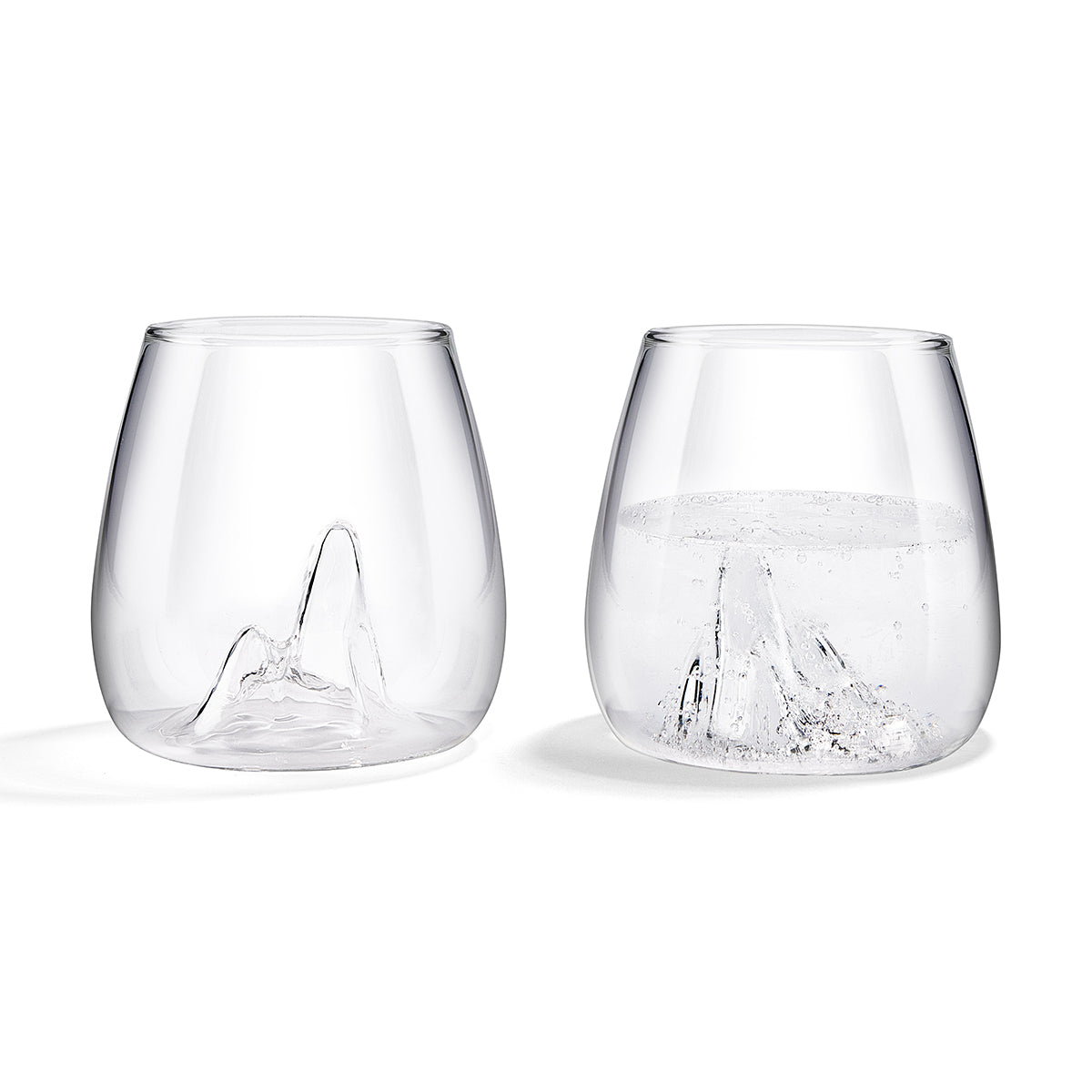 Image of a pair of glass tumblers with the shape of a mountain peak indented into their base. 1 glass is filled with sparling water. Set against a white background.