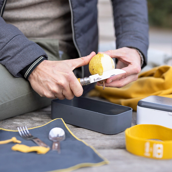 Person cutting a pear while using the outdoor lunch box filled with food and the cutlery handle and knife/spoon/fork kit and napkin on wooden picnic table 