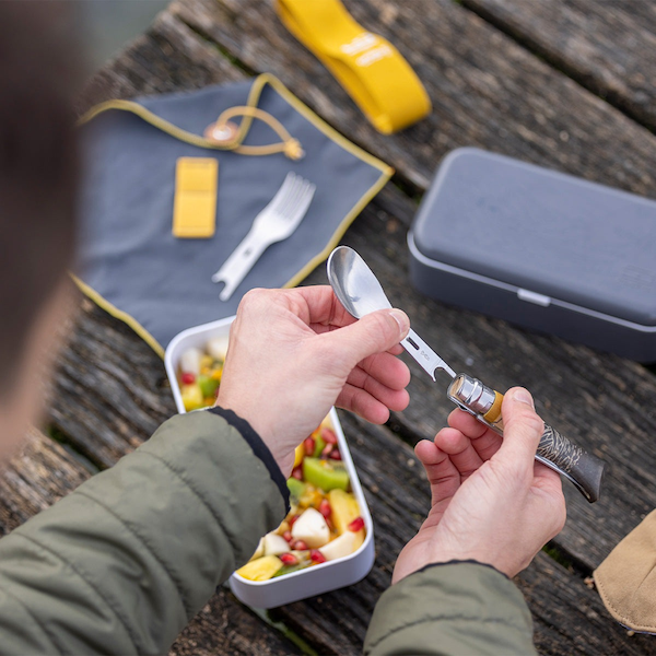 Man holding the spoon insert and cutlery hand above an outdoor lunch box filled with food and knife/spoon/fork kit and napkin on wooden picnic table insert & handle 