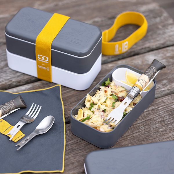 closed outdoor lunch box with 1 section filled with food and the cutlery handle and knife/spoon/fork kit and napkin on wooden picnic table 