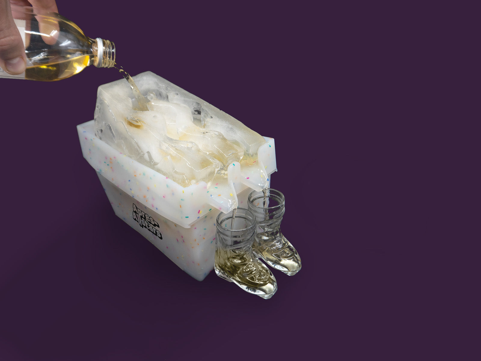 Photo of a white speckled plastic mould and miniature white plastic ski boot bindings, with a block of ice on top that has been molded in the shape of a slalom slope, with 2 funnels within the ice through which yellow liquor is poured to chill it. The yellow liquor runs through the ice and into waiting shots glasses shaped like ski boots. The image is set against a purple backdrop, viewed from the right hand side.