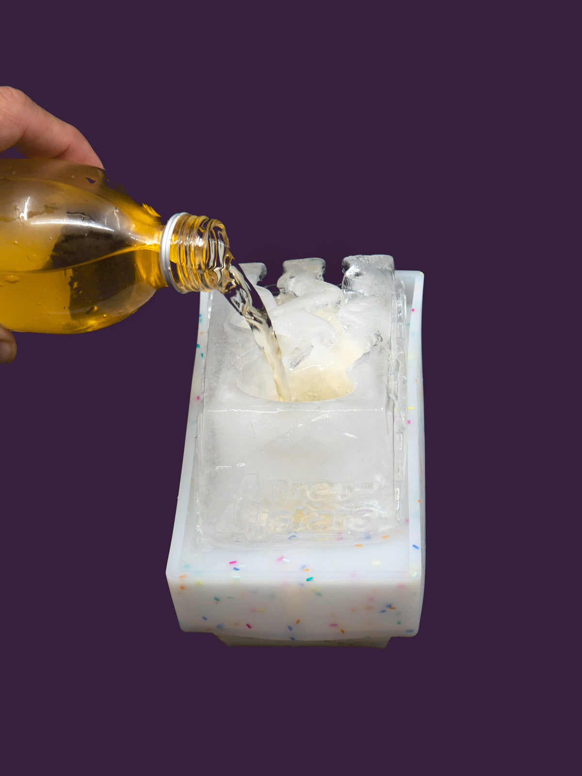 Photo of a white speckled plastic mould, with a block of ice on top that has been molded in the shape of a slalom slope, with 2 funnels within the ice through which yellow liquor is poured to chill it. The yellow liquor runs through the ice and into waiting shots glasses shaped like ski boots. The image is set against a purple backdrop, viewed from the back.