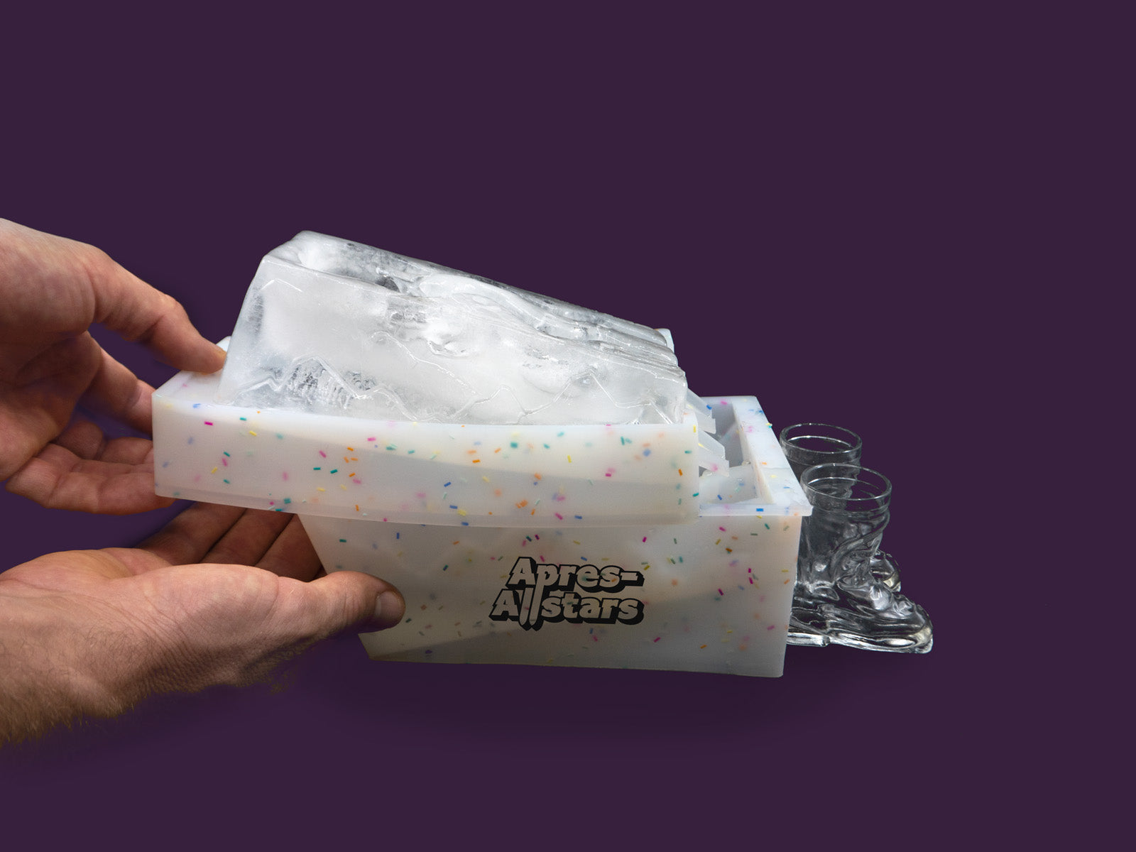 Photo of a white speckled plastic mould, with a block of ice on top that has been molded in the shape of a slalom slope, with 2 funnels within the ice through which liquor is poured into waiting shots glasses shaped like ski boots. The image is set against a purple backdrop, viewed from the right hand side. A pair of hands are moving the top and bottom of the plastic mold.