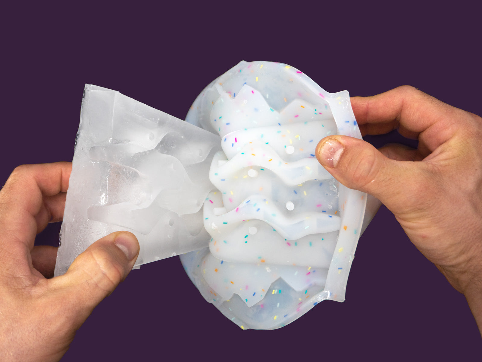 Photo of a white speckled plastic mould being peeled away from the frozen water inside it by a pair of hands.The image is set against a purple backdrop, viewed from the back.Photo of a white speckled plastic mould has been separated from the frozen water inside. 1 hand is holding the mold, the other the ice.The image is set against a purple backdrop, viewed from the back.