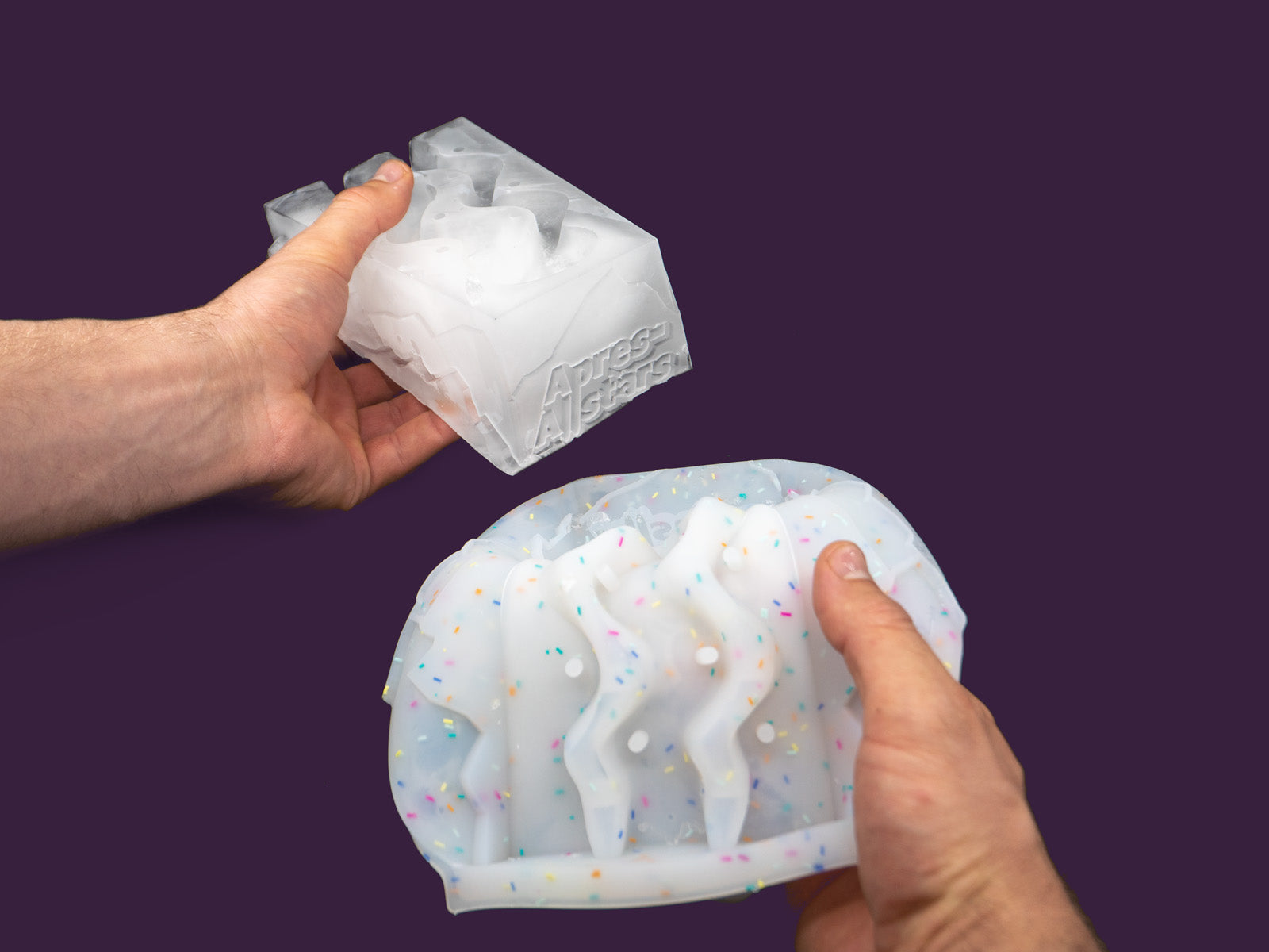 Photo of a white speckled plastic mould has been separated from the frozen water inside. 1 hand is holding the mold, the other the ice.The image is set against a purple backdrop, viewed from the back.