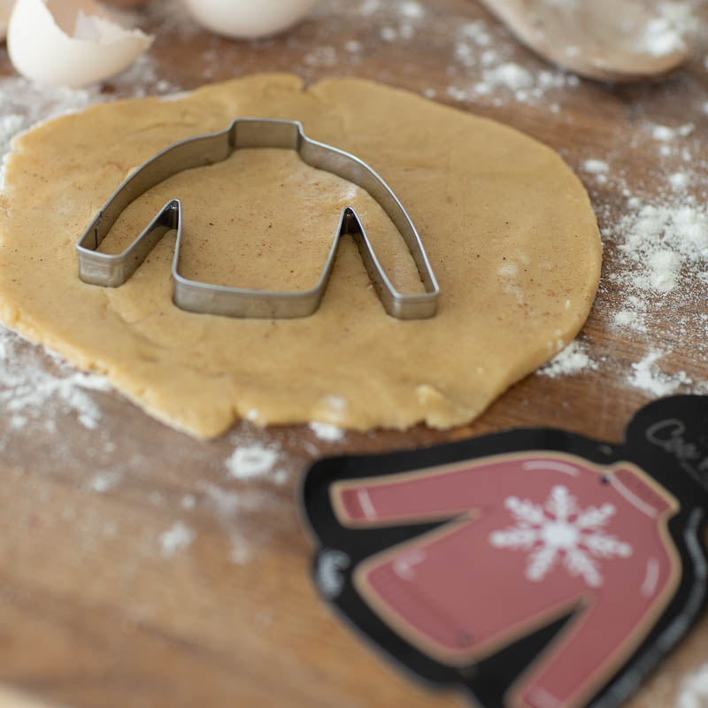 Image of jumper cookie cutter on wooden pastry board with pastry rolled out & the black cardboard cutter backing depicting a red jumper with a snowflake resting off to one side